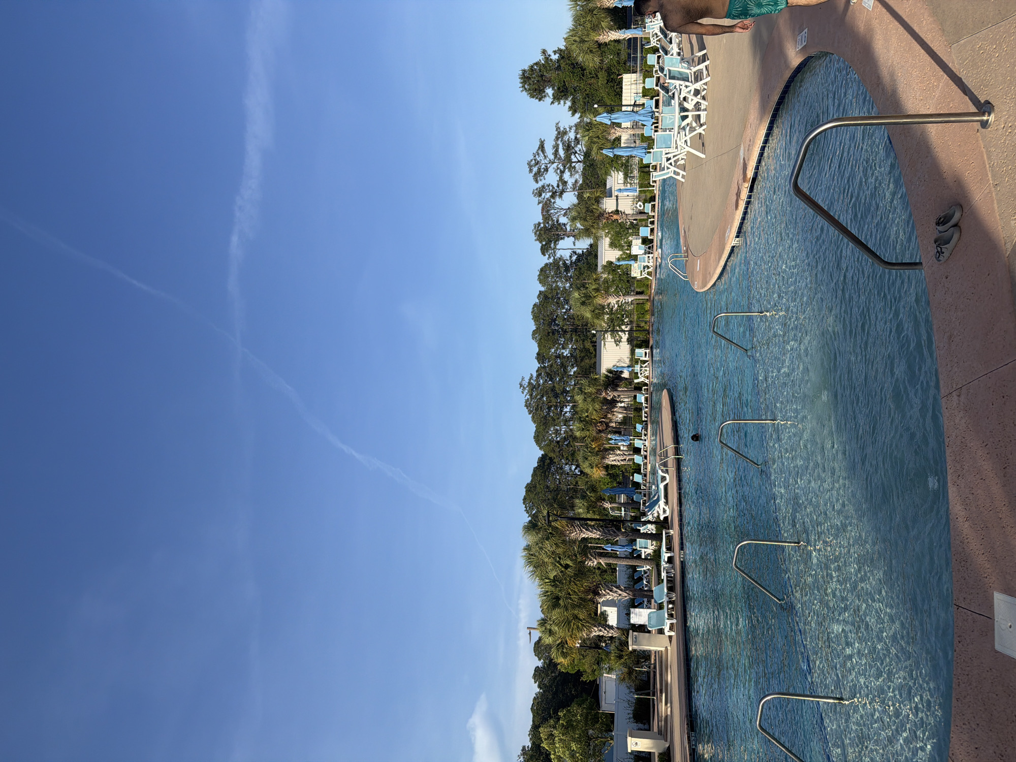 The image shows a swimming pool with lounge chairs and trees next to it. The sky is blue with some clouds. There is a person standing near the pool. The pool has handrails in the water. The location is Myrtle Beach, United States of America.