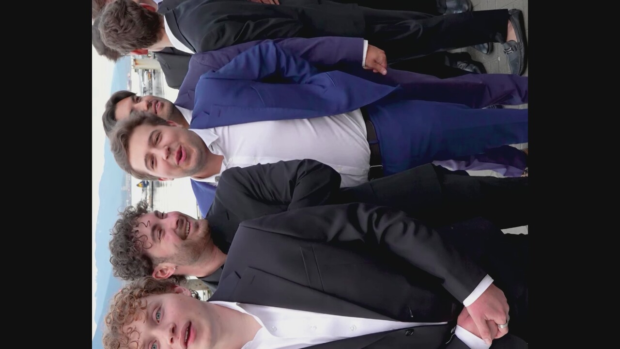 In Vancouver, Canada, a group of young men in formal attire, likely suits, are gathered outdoors. The foreground is dominated by three individuals. The person on the left, facing forward, has curly blonde hair and is wearing a black suit jacket over a white dress shirt. Next to him, a man with curly dark hair and a beard is looking to his right and smiling, also in a black suit. The central figure is wearing a vibrant blue suit and a white shirt, with his mouth open as if speaking, and is looking towards the viewer. Behind them, other men in suits are visible, suggesting a formal occasion. The background depicts a body of water with boats and hills in the distance, characteristic of Vancouver's coastal scenery. The ground appears to be paved, possibly a boardwalk or pier.