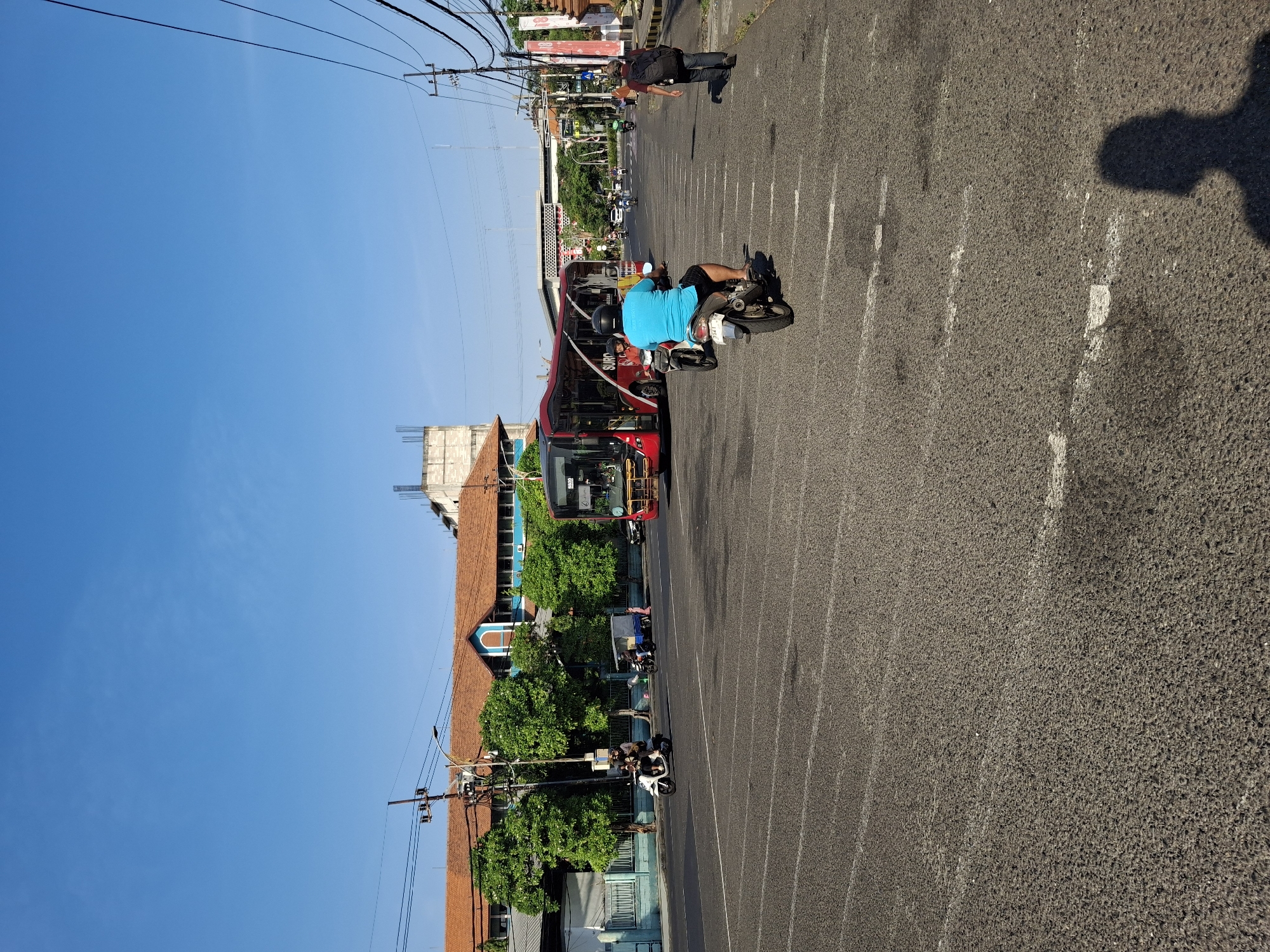 The image depicts a street scene in Surabaya, Indonesia, under a clear blue sky. A red and white bus, identified by the text "TRANS SURABAYA" on its side, is prominently visible in the mid-ground, facing left. Several motorcycles are in motion on the asphalt road, which is marked with white lines. In the foreground, a person on a motorcycle wearing a blue shirt is seen from behind, facing away from the camera. To the right, another person with a backpack walks away from the camera. Buildings with terracotta roofs and a construction site are visible in the background. Trees line the side of the road. The overall impression is of a typical urban street during daylight hours.