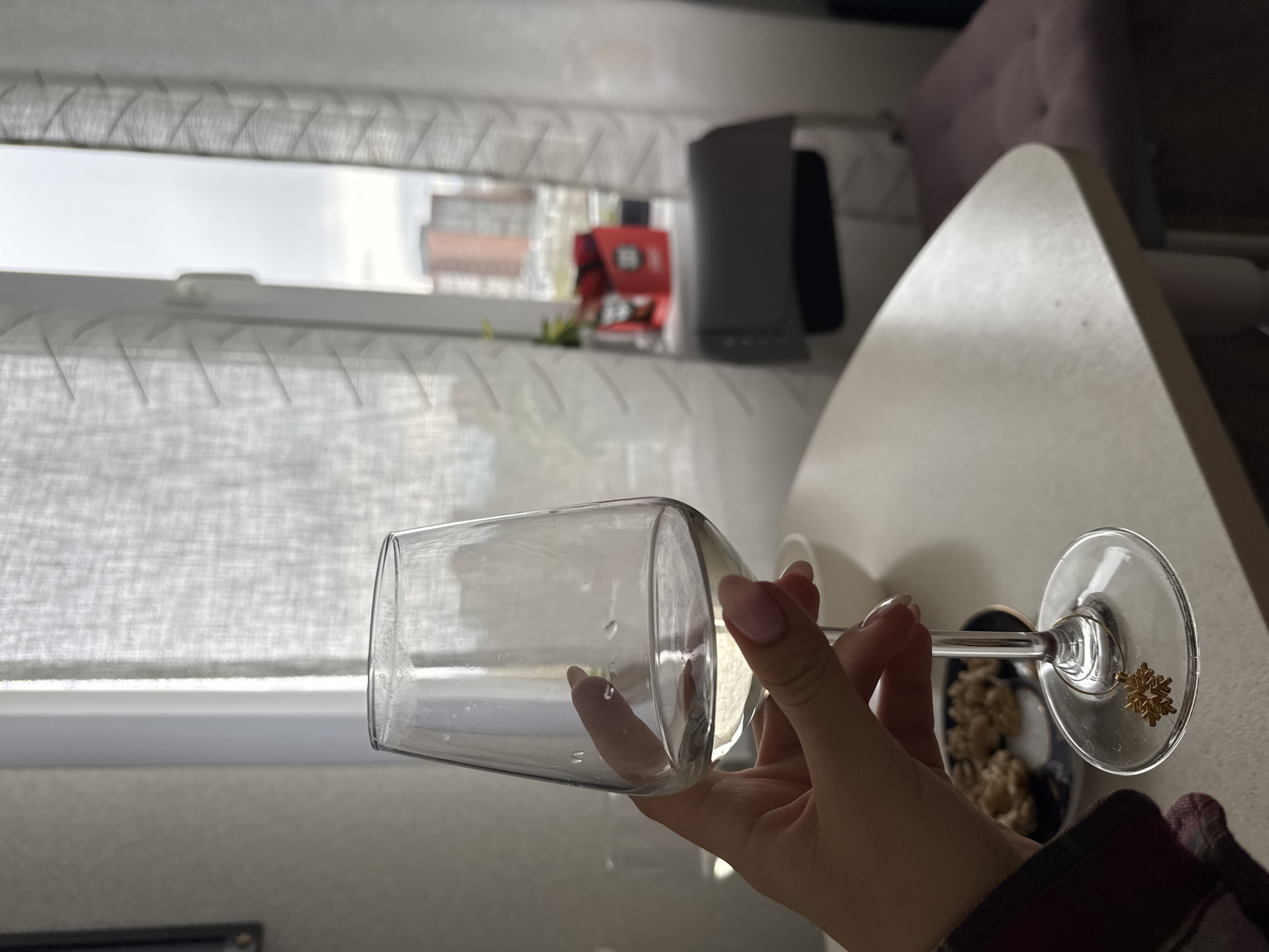 A close-up, slightly tilted image captures a hand holding a clear, empty wine glass with a delicate stem. The glass shows droplets of liquid on its interior surface, suggesting it has recently been used or rinsed. A small, gold-colored snowflake charm is attached to the stem near the base. The hand, with neatly manicured light pink nails, grips the stem of the glass.
In the background, a large window is visible, covered by translucent white blinds or curtains that diffuse the light. Beyond the window, an overcast sky and blurred outlines of distant buildings can be discerned. On the windowsill, a small red object with unidentifiable white markings and a dark gray rectangular device are placed. To the right of the frame, part of a light-colored, curved table surface is visible, along with a blurred pink upholstered item, possibly a headboard or cushion. A bowl containing what appears to be nuts or cereal is partially visible behind the glass. A sleeve of plaid fabric, likely from a shirt or robe, is seen in the lower right corner. The scene appears to be an indoor setting during the daytime.