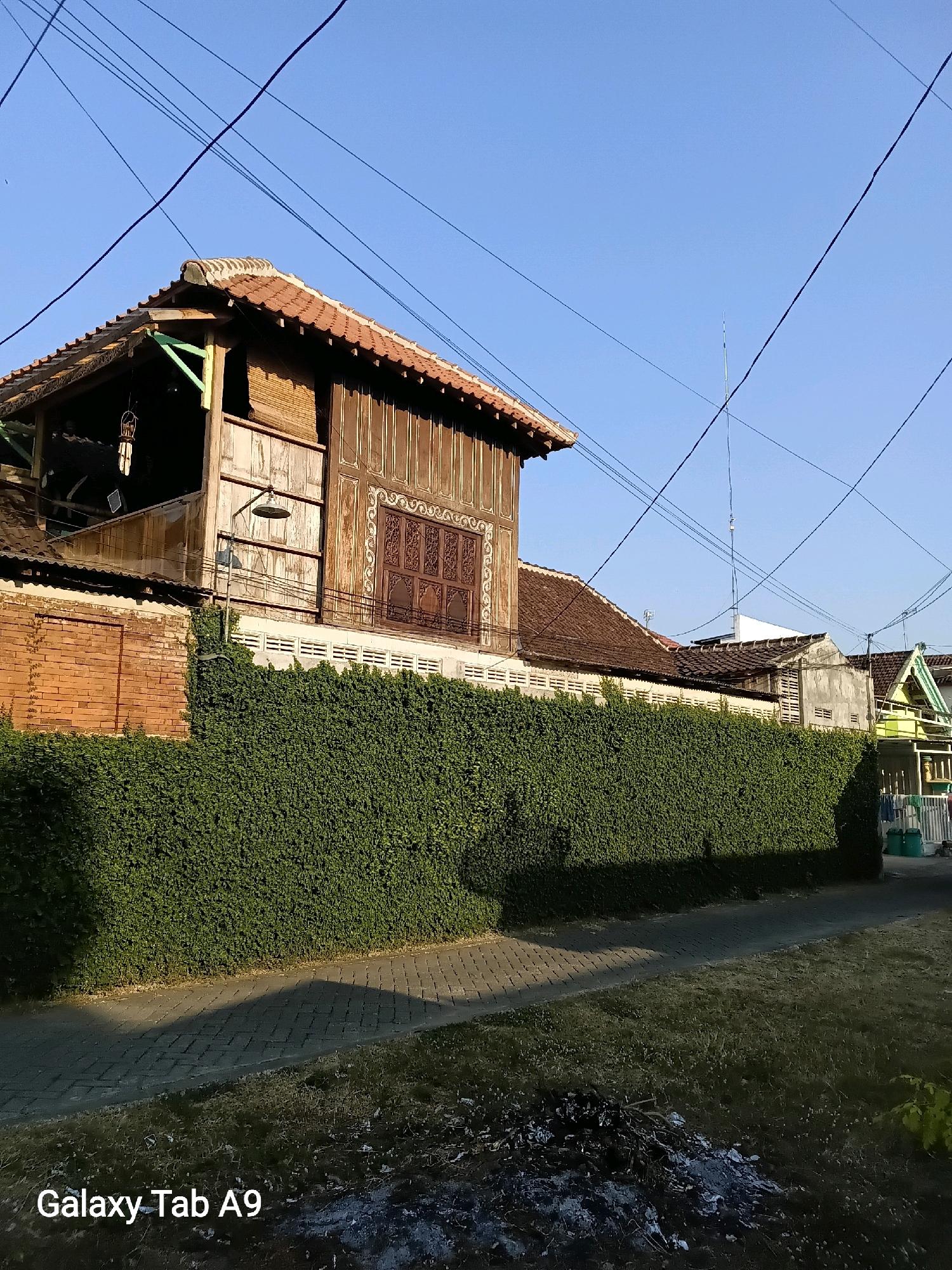 The image depicts a residential street scene in Kelutan, Indonesia, bathed in the warm glow of sunlight under a clear blue sky. The foreground features a paved walkway with a shadow cast by a dense, well-maintained green hedge that runs along the property line. Beyond the hedge, a traditional Indonesian house with a weathered wooden facade and a terracotta tile roof is visible. The house has intricate wooden carvings on its windows and an open upper story. Adjacent to it are other houses with similar architectural styles. Overhead, a network of power lines crisscrosses the sky. In the lower right corner, a small pile of burnt debris is present on the grassy area. The overall impression is one of a quiet, established neighborhood with a blend of traditional and modern elements.
