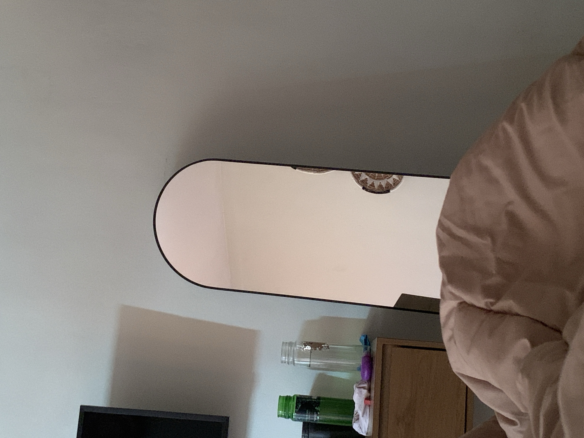 A horizontal, slightly low-angle shot presents a portion of an interior setting. A long, rounded-edge mirror with a black frame hangs on a light grey wall, reflecting the room's ceiling, which features a woven decorative plate. Beneath the mirror are a collection of items, including a black television screen, two glass bottles (one clear with silver contents and the other green), and a wooden dresser. A partially visible, large, light pink fluffy object dominates the right side of the frame. The scene appears to be a domestic interior. The location in Kuta Selatan, Indonesia, is not discernible from this image.