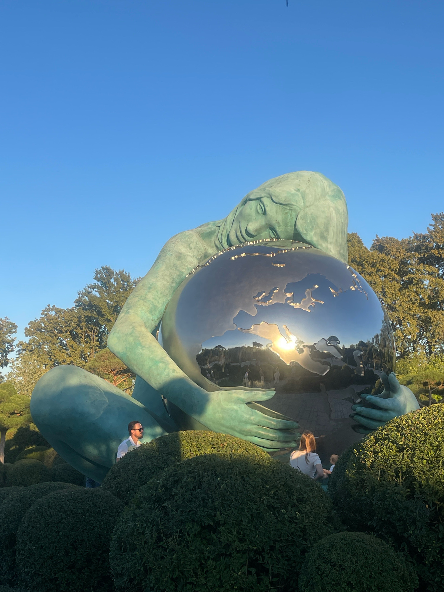 The image shows a large, green sculpture of a reclining female figure holding a reflective sphere, which appears to be a model of the Earth, in her arms. The sculpture is situated outdoors, with a clear blue sky in the background and lush greenery surrounding its base. Several people are visible in the foreground, appearing to be visitors interacting with the artwork. The sun is setting, casting a warm glow and creating reflections on the sphere. The overall scene suggests a public park or art installation. The location is the "Skypark" in Krasnodar, Russia, where this sculpture is a prominent feature.