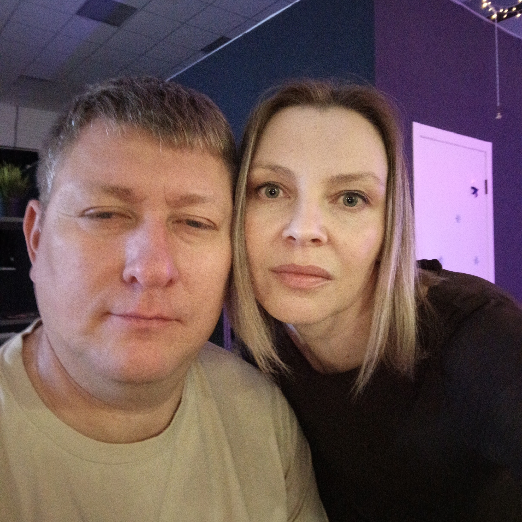 A photo depicts a middle-aged man and a middle-aged woman. The man has short, graying hair and is wearing a light-colored t-shirt. He appears to be squinting in one eye. The woman has shoulder-length blonde hair and is wearing a dark shirt. The background consists of a dark blue wall, a white door with a small sign, and ceiling lights. The location is possibly inside a building in the city of Chaikovsky, Russia, but without further information, that cannot be definitively confirmed.