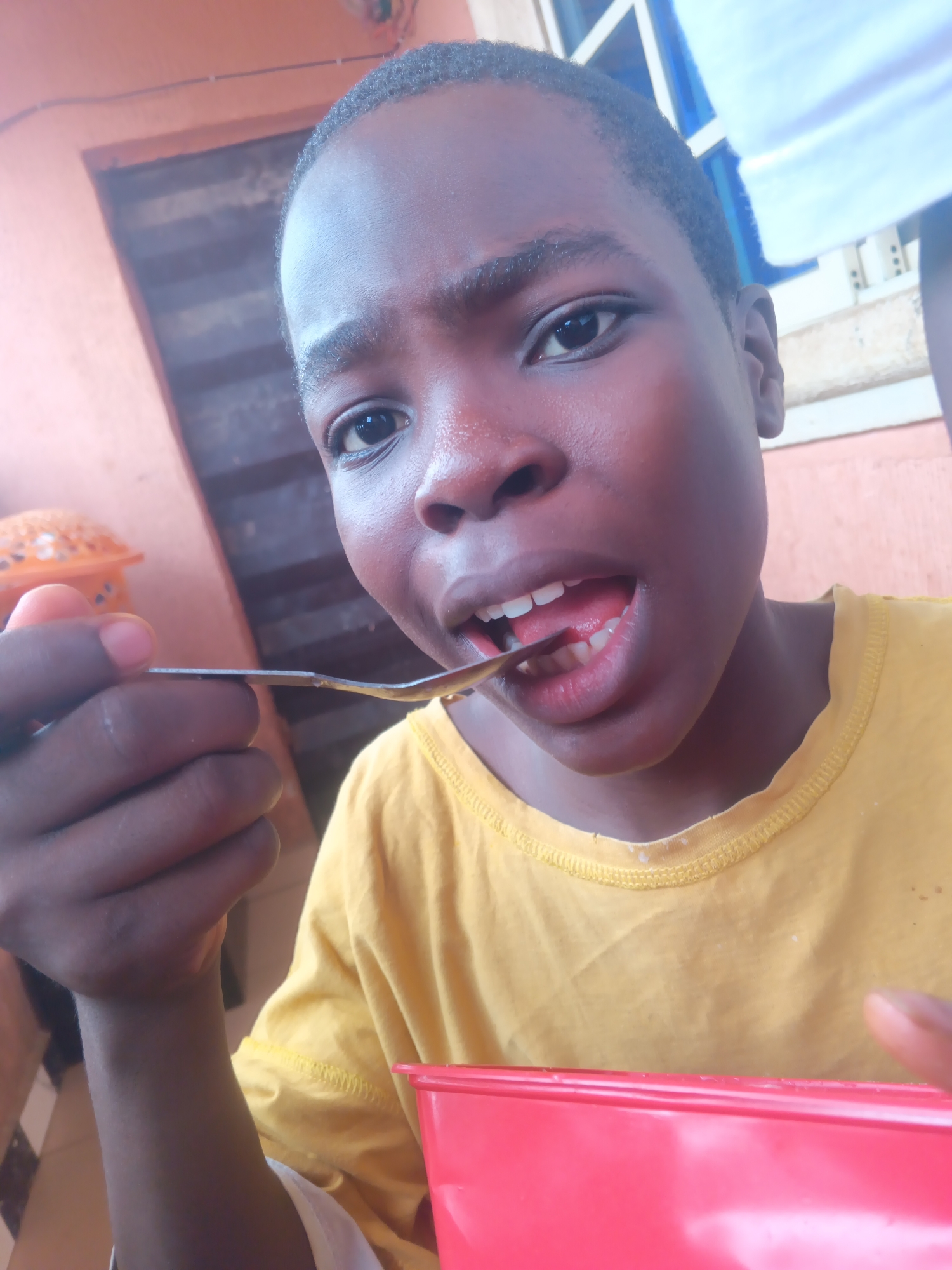 A young African boy is eating from a red bowl with a spoon. He is wearing a yellow t-shirt, and his face is shiny with perspiration. The location is Onitsha, Nigeria, as evidenced by the architecture in the background and the individual's ethnicity. The background shows a house interior and a window. The boy appears to be indoors.
