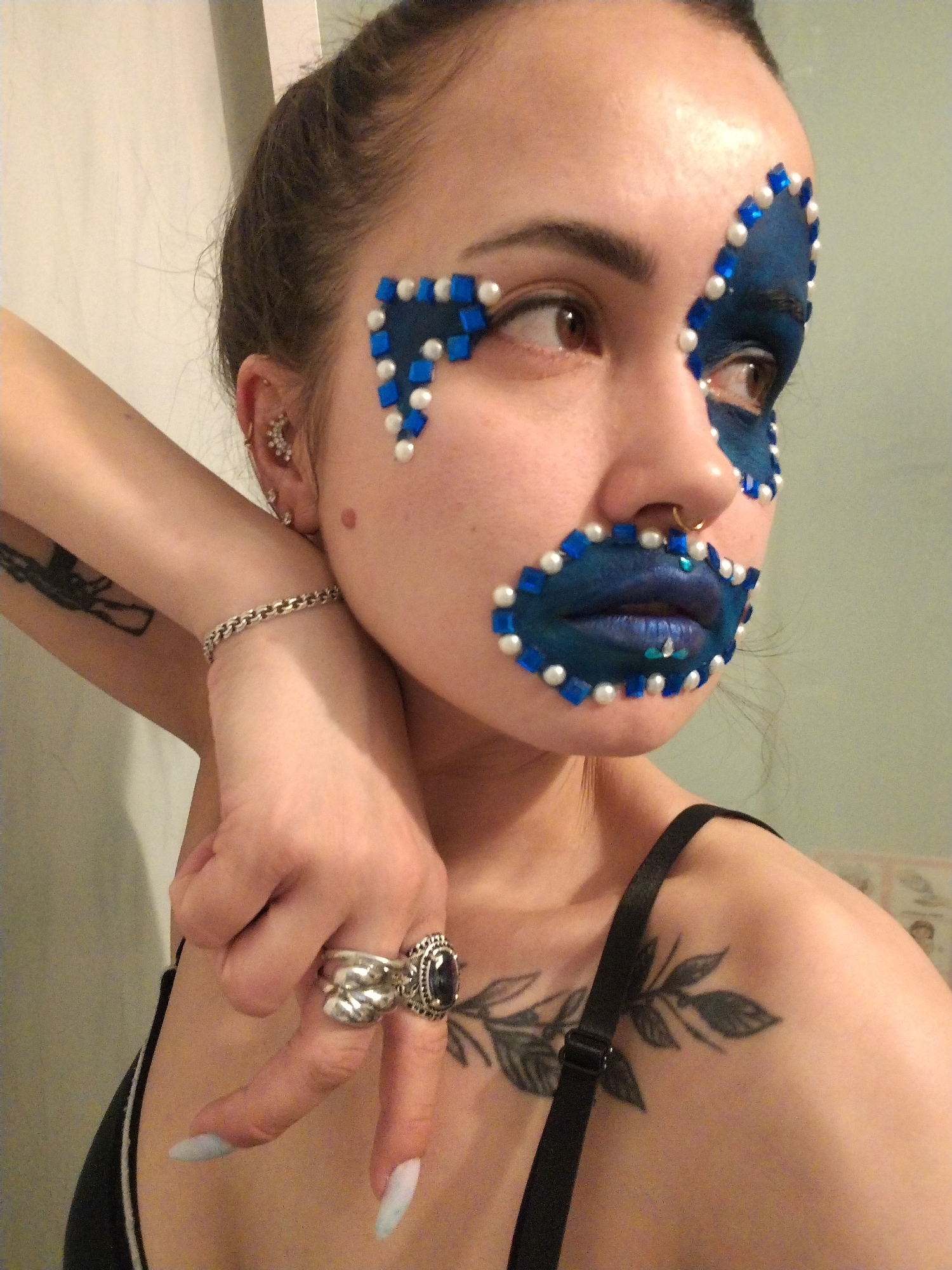 A young woman with elaborate face art is captured in a close-up, chest-up portrait indoors. Her face is adorned with dark blue eyeshadow and lipstick, outlined and accented with an array of blue square rhinestones and white pearl-like beads, creating a mask-like effect around her eyes and mouth. Her left eye is visible, featuring brown iris, and her lips are painted a shimmery blue. She has a nose ring (septum piercing), multiple ear piercings on her left ear (including a cluster earring and a barbell), a silver chain bracelet on her left wrist, and two rings on her right hand – a large silver ring with a dark oval stone and a smaller, ornate silver ring. Her nails are long, pointed, and light blue. A black bra strap is visible on her right shoulder, and she has a tattoo of a leafy branch on her right collarbone area, and another dark tattoo on her left upper arm. She is posing with her left arm bent, her right hand holding her left forearm near her neck. The background is a simple, light-colored wall, suggesting an indoor setting, possibly in Noviy, Russia. There is no visible text in the image. The lighting appears to be artificial, making the time of day undeterminable.