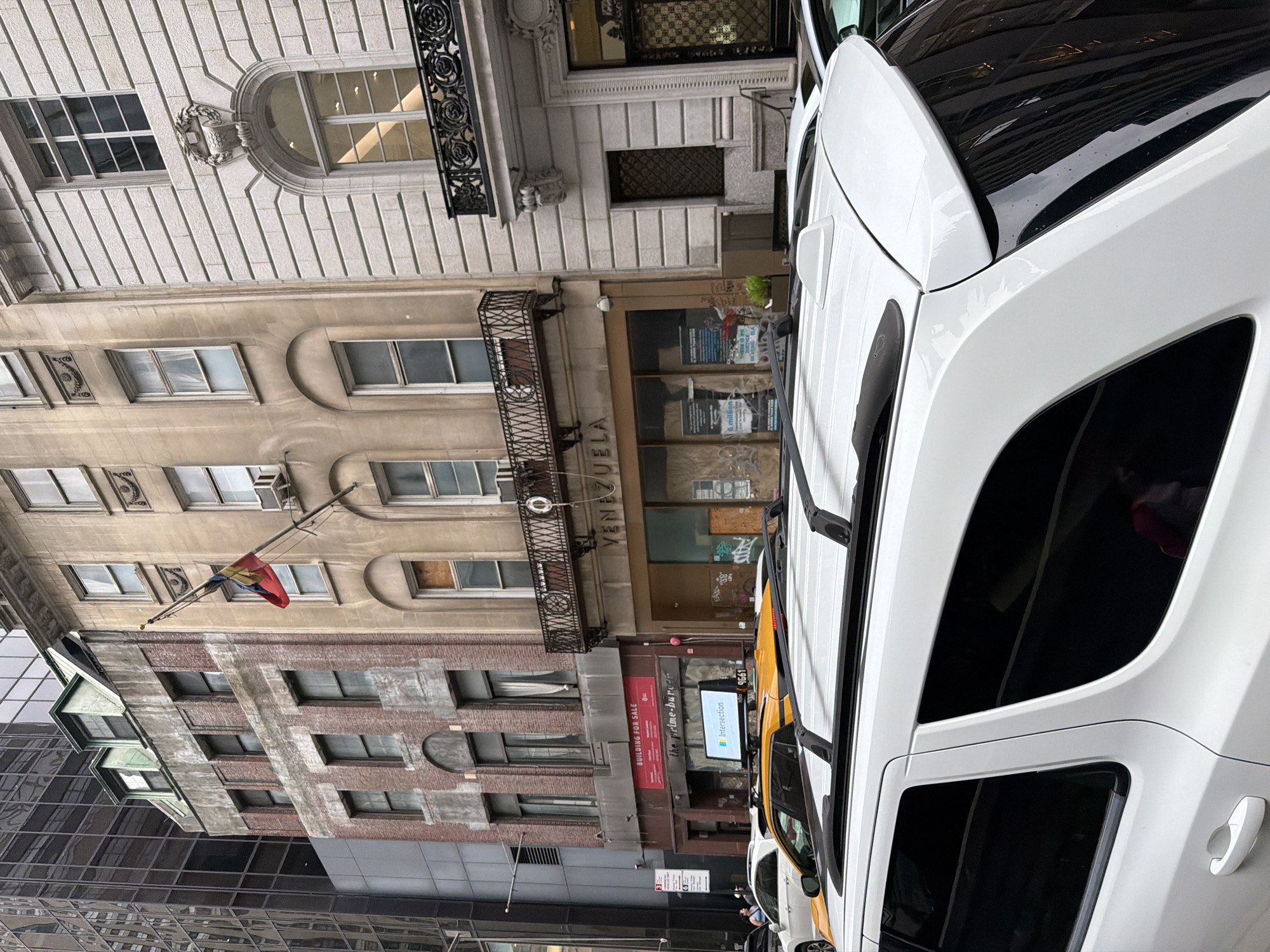 The image captures a street scene in New York City, United States of America. The foreground is dominated by the side of a white SUV, partially obscuring the view. Behind the SUV, several buildings line the street. The architecture includes brick and stone structures with windows, some of which have boarded-up sections. A Venezuelan flag is visible, attached to one of the buildings. Signage reading "VENEZUELA" is visible above a building entrance. Other commercial signs are present but their full details are partially obscured. Traffic, including a yellow taxi, is visible. The environment suggests an urban setting with a mix of architectural styles and street-level commercial activity.