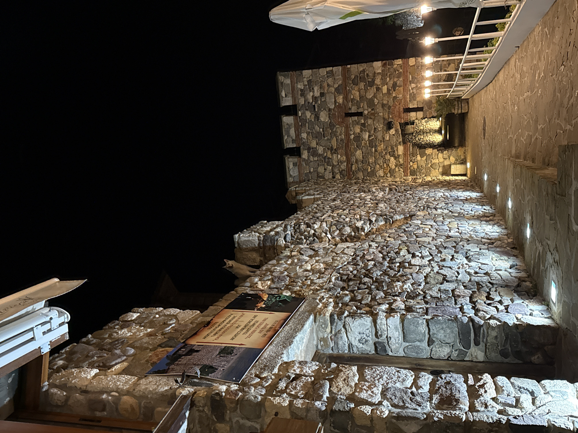 The image depicts an illuminated stone architectural complex at night in Sozopol, Bulgaria. The central focus is a prominent stone wall on the left, constructed from irregular light-colored stones with visible mortar. Near the top of this wall, a light-colored cat with pointed ears stands looking towards the viewer. Attached to the wall, approximately one-third of the way from the left edge, is a rectangular sign with both Cyrillic and Latin text. The Latin text reads: "VISIT ARCHITECTURAL HISTORICAL AND TOURIST COMPLEX 'SOUTHERN FORTRESS WALL AND TOWER' GSM: +359 899 34 74 14," confirming the specific location.
To the right of the initial stone wall, a paved stone pathway extends into the distance, illuminated by small, square, recessed ground lights and multiple street lamps mounted on white poles, featuring spherical lights. A white railing runs along the right side of the pathway. Further down the path, an archway leads to a darker interior, and beyond the railing, a large, folded white umbrella is visible. Another substantial stone structure, possibly a tower or a section of a fortress, is positioned in the mid-ground, constructed from similar irregular stones with horizontal bands of red brick visible, and featuring crenellated battlements at its top. The sky above is entirely dark, indicating nighttime conditions. Artificial warm and cool lighting casts strong shadows and highlights on the textured stone surfaces.
