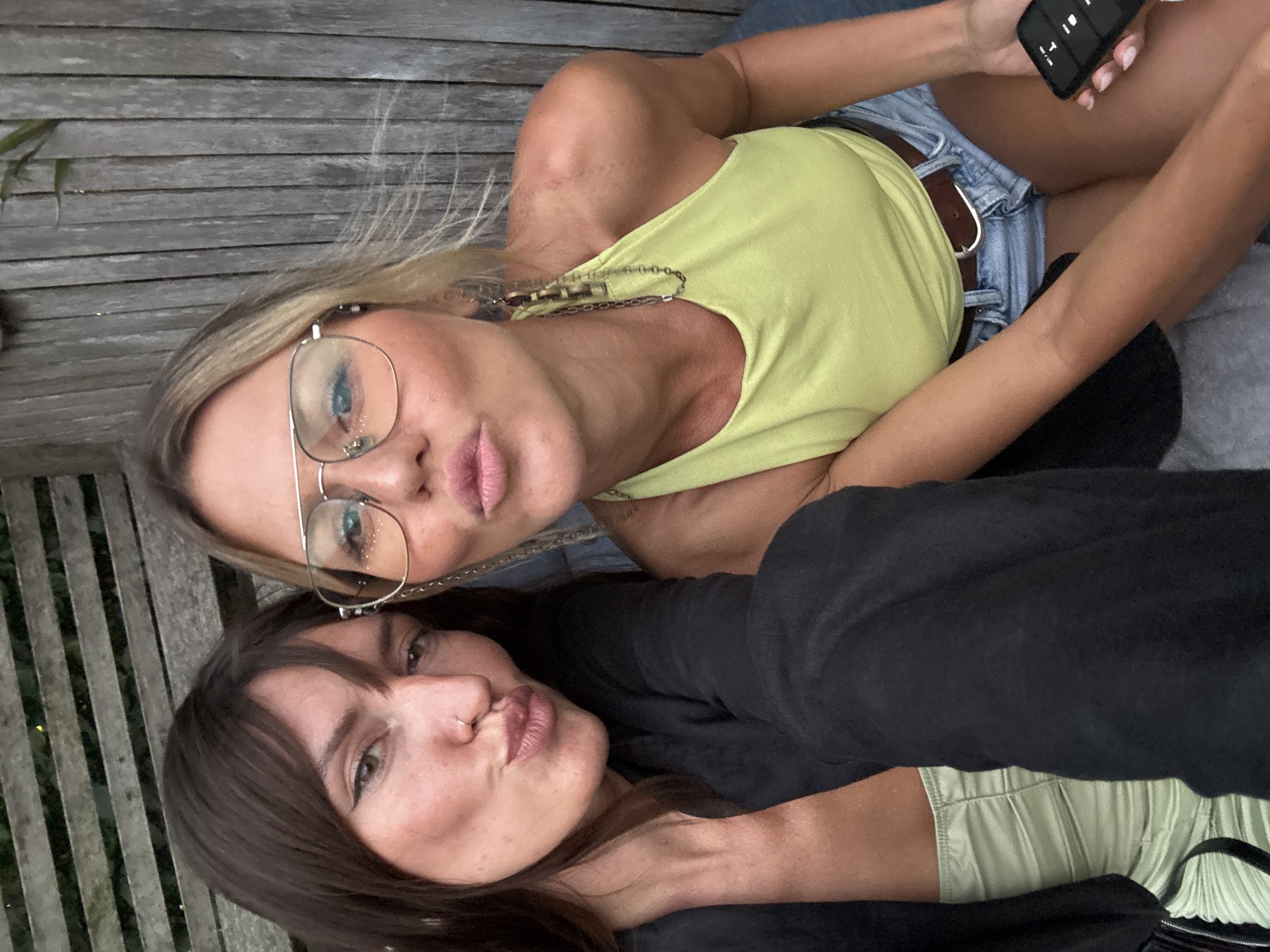 Two women are taking a selfie outdoors in front of a wooden fence. The woman on the left has long dark hair and is wearing a black jacket over a light green ruched top. The woman on the right has blonde hair, is wearing oversized glasses, a bright yellow tank top, and denim shorts. She is holding a smartphone in her right hand. Both women are puckering their lips as if for a kiss. The background consists of a weathered wooden fence and some foliage. The lighting suggests it is daytime, likely overcast or late afternoon.