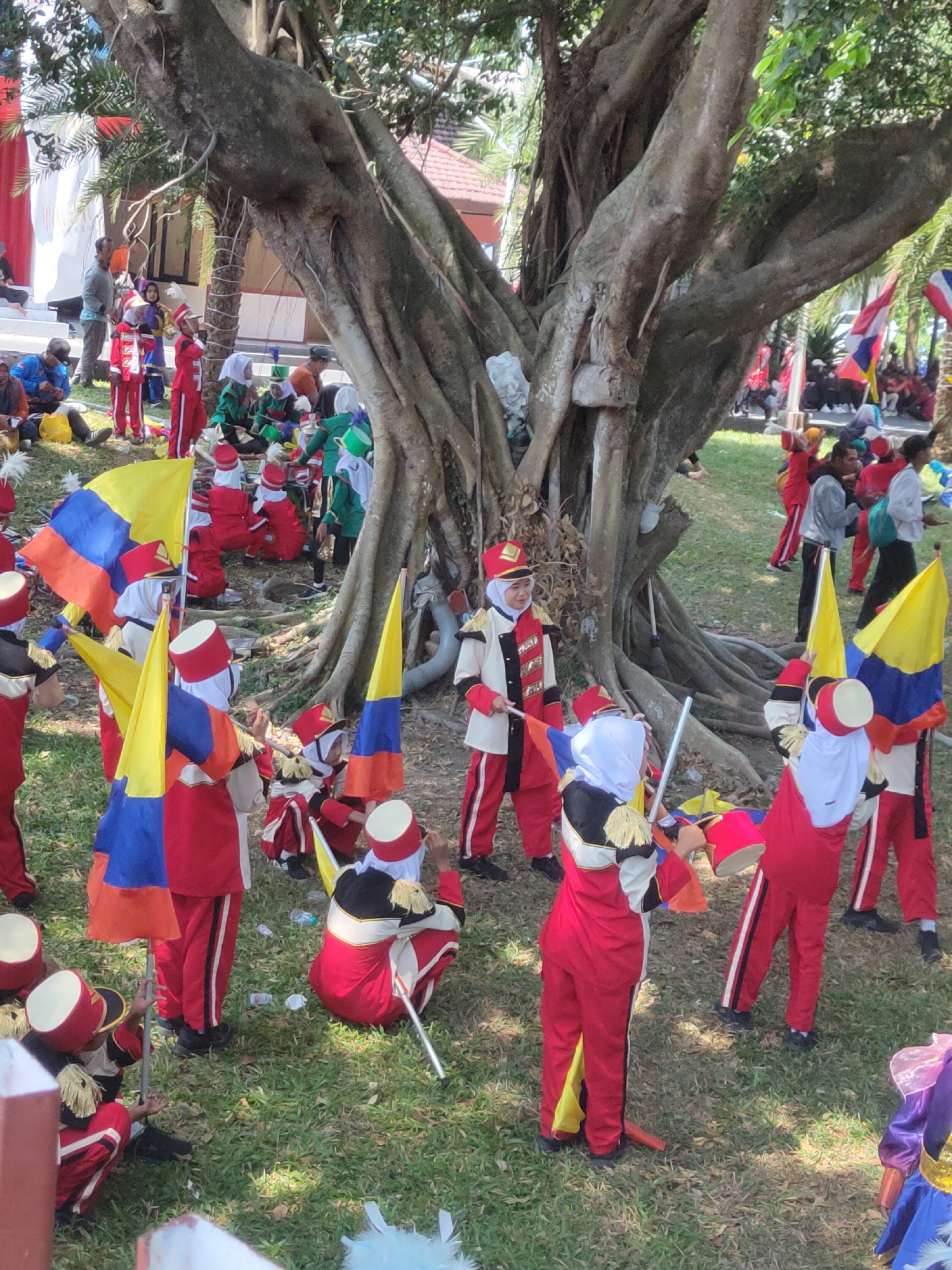 The image depicts a group of children dressed in marching band uniforms, likely participating in an event at the Alun-Alun Ngasem in Kediri, East Java, Indonesia. The children, mostly wearing red and gold uniforms with white headscarves or hats, are holding flags and standing on a grassy area under the shade of a large tree with exposed roots. Some children are seated or kneeling. In the background, other individuals, some also in uniform, are visible, along with buildings and trees. The atmosphere suggests a community gathering or performance.