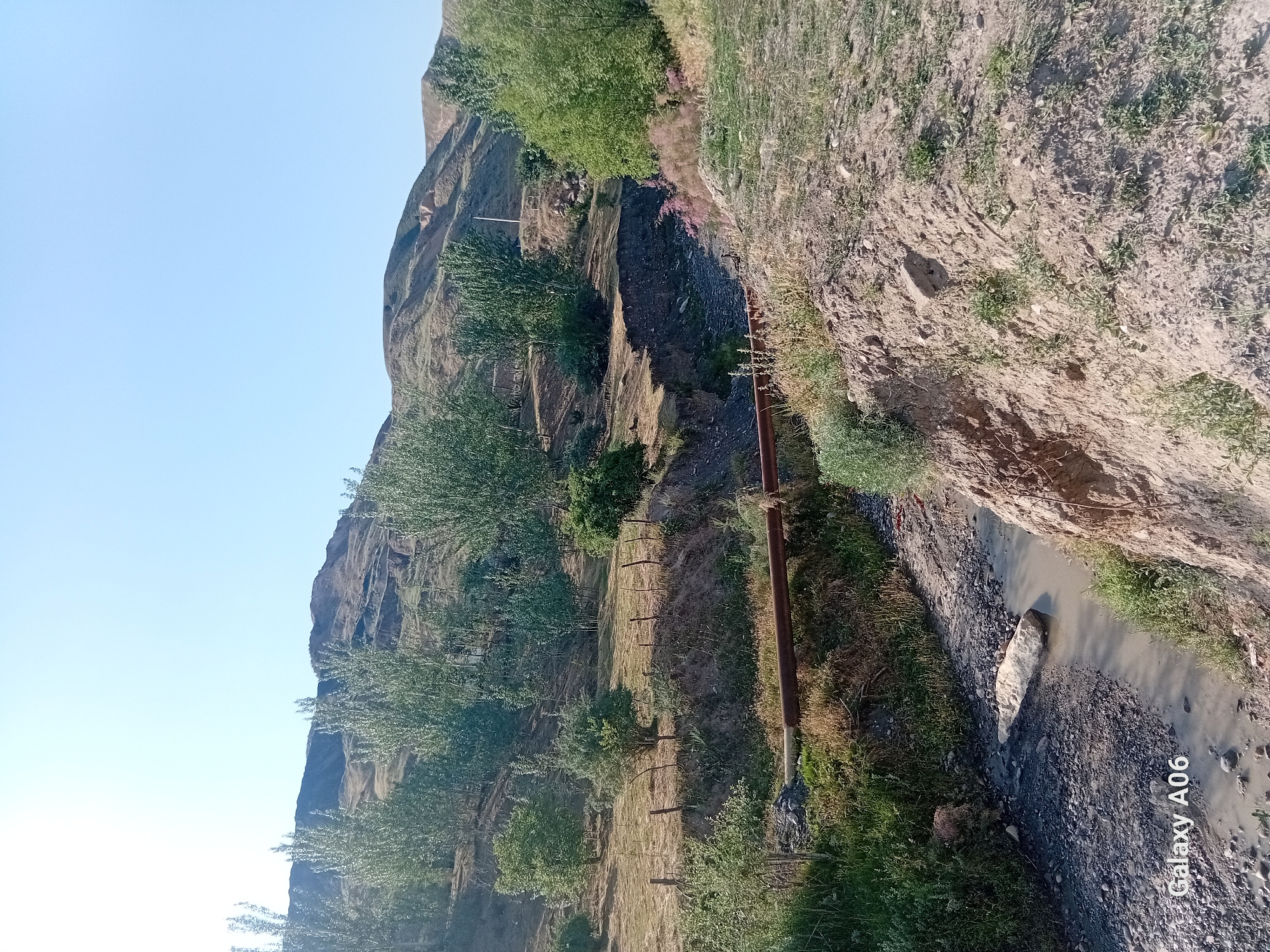 A wide-angle, low-angle outdoor shot depicts a dry, rocky stream bed with a shallow, murky stream flowing through it. The stream bed is lined with dark grey rocks and gravel, with some sparse green vegetation growing on its banks. A large, light-colored rock is partially submerged in the stream. A rusty metal pipe runs horizontally across the stream bed. The background features rolling hills covered with dry grass and scattered trees. The sky is clear and blue. In the bottom left corner, a watermark reads "Galaxy A06". The scene appears to be located in Kojo-Aryk, Kyrgyzstan.