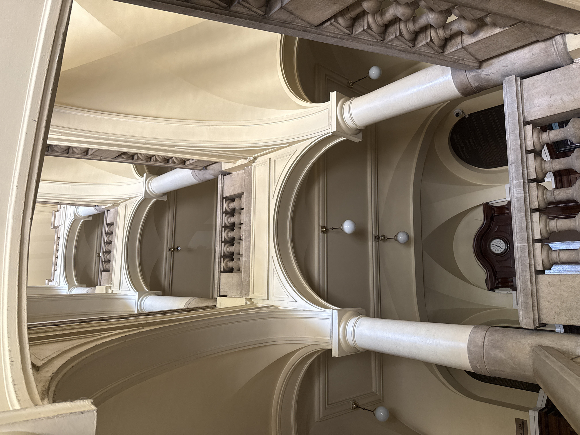 The image displays a grand and ornate interior, likely within a historic public building or a lavish residence, viewed from a low angle looking upwards into a vast, curvilinear space. This architectural structure strongly suggests a spiraling stairwell or an elaborate atrium that extends upwards through multiple floors.
The dominant color palette consists of warm off-white, cream, and light beige tones covering the vaulted ceilings and walls. These light colors contrast with darker elements of carved stone and wood used for decorative details. The architecture is rich in classical influences, featuring numerous graceful arches that ascend, supported by sturdy, smooth columns with elegantly designed capitals and bases. Ornate stone balustrades, complete with finely carved balusters and decorative moldings, delineate the upper levels, indicating the presence of multiple floors. A dark, intricately carved wooden frieze or cornice, adorned with repeating architectural motifs, crowns the upper sections of the structure.
Several spherical, white light fixtures are mounted on the walls, providing ambient illumination. On one of the upper levels, a circular clock with Roman numerals is set within a dark, possibly wooden, panel, indicating the time to be approximately 1:55. A small, dark security camera is subtly visible near one of the columns.
A recessed, oval-shaped panel on an upper wall features partially obscured text written in Cyrillic. discernible words include "ЦЕНТРАЛЕН" (Central), "ИЗТОК" (East), "ПОЛУЕТАЖ" (Mezzanine), "ПРОХОД" (Passage), and "ЛИФТ" (Elevator), suggesting a directional sign or building directory. A smaller sign, "TA - 136", is visible in the lower right portion of the image.
The absence of people emphasizes the architectural grandeur and the historical ambiance of the space. The overall impression is one of elegance, history, and quiet majesty, characteristic of a significant cultural or governmental building in a European city, consistent with the location context of Sofia, Bulgaria.