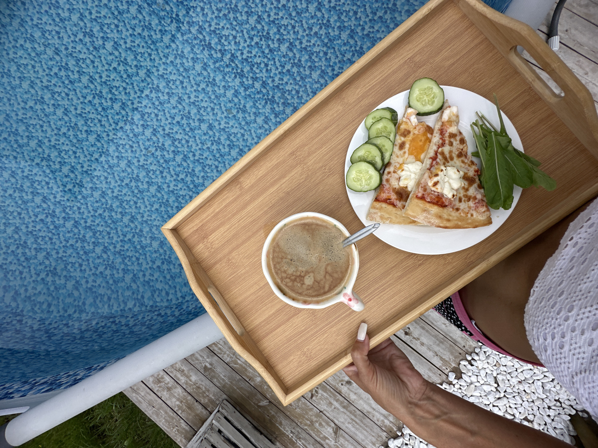 A wooden tray with breakfast is held over a tiled swimming pool. On the tray are two slices of pizza, sliced cucumbers, a small bunch of greens, and a mug of coffee with a spoon. A person's arm, with manicured nails, is visible holding the tray. The background consists of a blue tiled pool and wooden decking. The scene is outdoors.