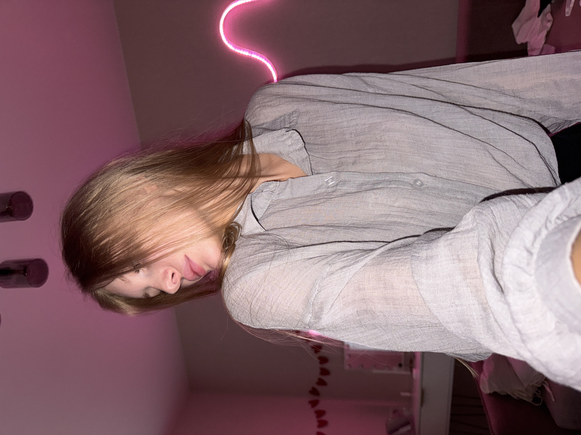The media file shows a young woman lying down, her head resting on her arm. She has long, blonde hair and appears to be wearing a light gray, long-sleeved shirt. Her eyes are closed, and her lips are slightly parted, suggesting a relaxed or sleepy expression. She is illuminated by a bright pink neon light, which curves behind her head and casts a vibrant glow on the wall and her clothing.
The background is dimly lit, with a soft pink hue on the wall. There are some cylindrical objects on the left side of the frame, and a hint of a shelf or dresser with some items on it in the upper right corner. A string of small heart-shaped lights is visible on the wall in the lower right portion of the image.
The overall mood is intimate and serene, enhanced by the warm, ambient lighting. The image appears to have been taken indoors, likely in a bedroom or personal space, given the decor and soft lighting. There is no visible text in the media file. The time of day is not specified, but the artificial lighting suggests it could be evening or night. The location context of Perm, Russia, provides no additional visual cues within the image itself.