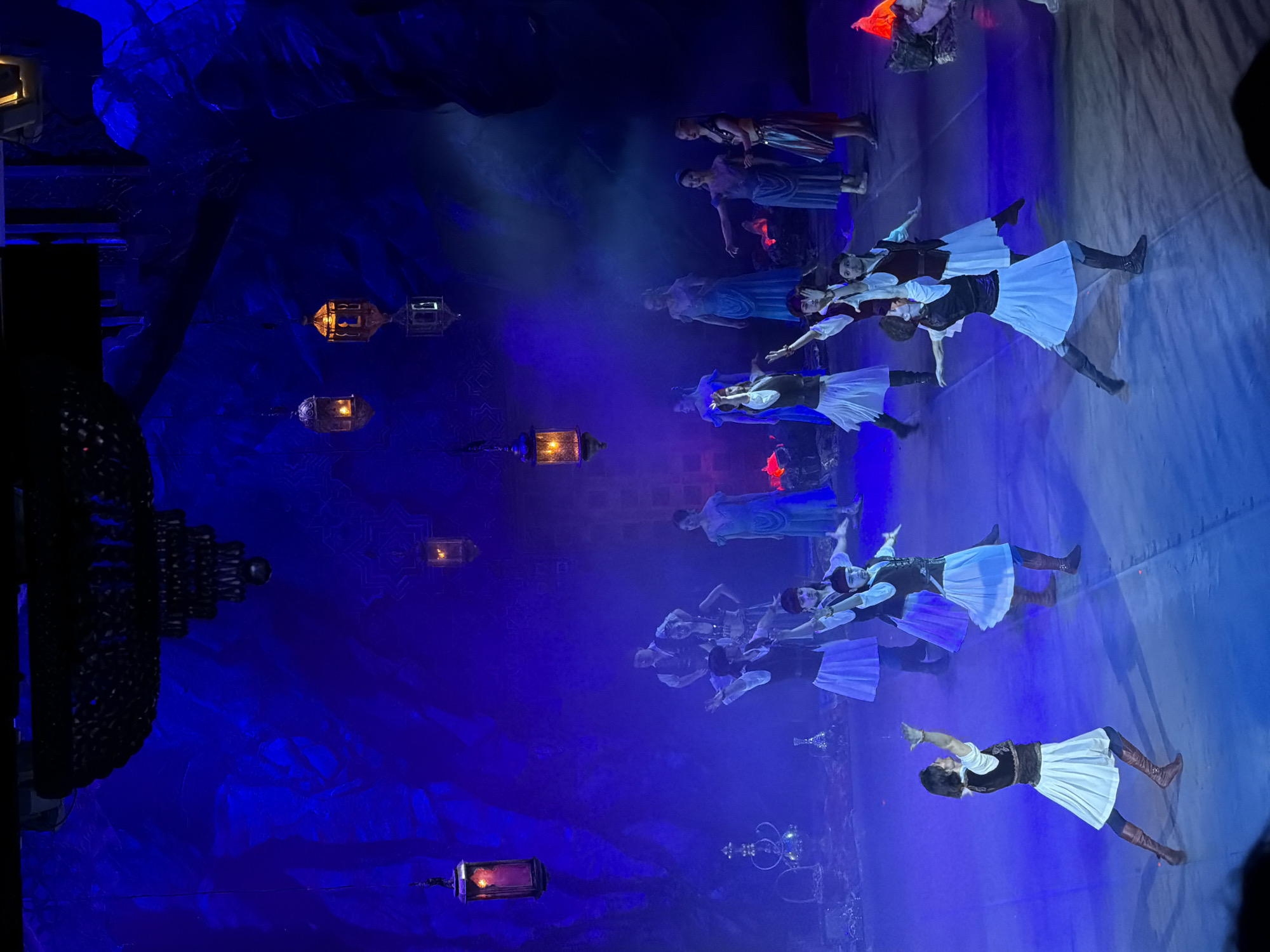 A medium-angle shot depicts a stage performance, likely in Izhevsk, Russia, given the architectural motifs and costuming styles commonly associated with that region. The scene is bathed in cool, blue light, enhancing the atmospheric feel. A group of dancers, dressed in white skirts, dark vests, and high boots, are posed dynamically, seemingly mid-performance. Their arms are extended, contributing to a sense of movement and grace. Additional figures, further back on the stage, are clad in flowing blue gowns, adding depth to the composition. Suspended in the background are decorative lanterns that cast warm light. The backdrop appears to be a wall adorned with intricate, possibly Arabian-inspired designs. The overall impression is of a staged, artistic performance, emphasizing the blend of cultural elements in Izhevsk's performing arts scene.