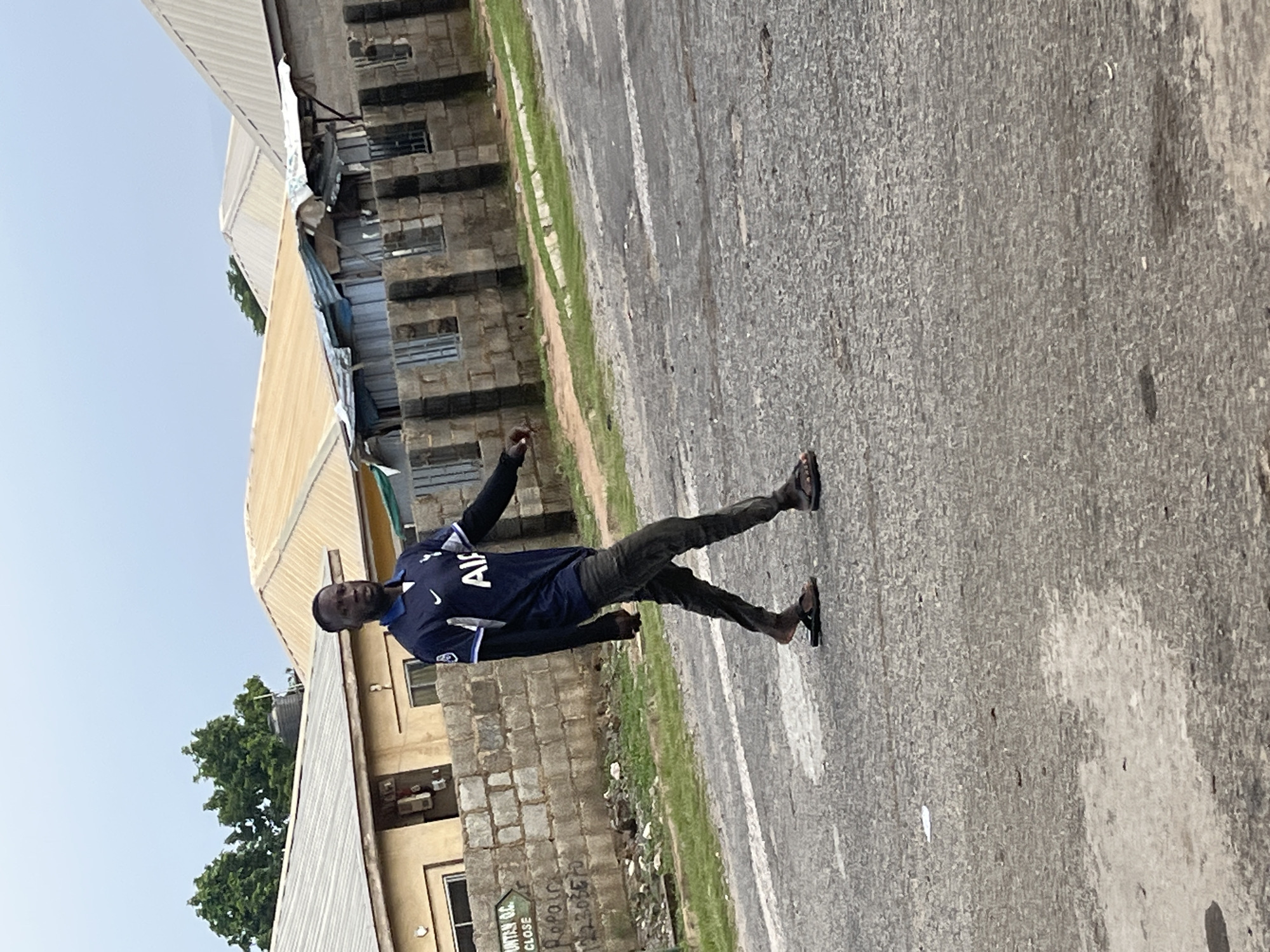 A Black man wearing a dark blue soccer jersey with white lettering that appears to say "AIA" and white trim around the collar and sleeves, is walking across a paved road. He is wearing dark, fitted pants and dark open-toed sandals. He is facing left and looking forward. Behind him, to his left, is a low stone wall. Beyond the wall are buildings with corrugated metal roofs and concrete block construction. One building has a beige facade. There is some green vegetation growing near the wall and on the side of the road. The sky is overcast. The scene appears to be in an urban or semi-urban area of Abuja, Nigeria, possibly a residential or commercial district.