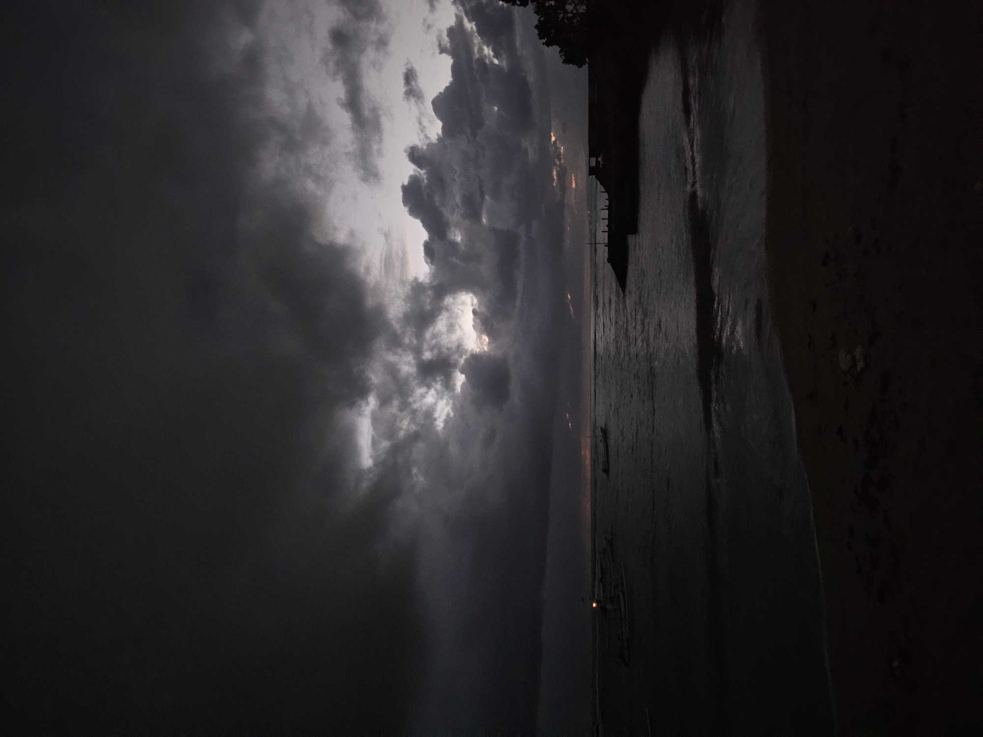 This is an image of a beach at dusk near Denpasar, Indonesia. The sky is heavily overcast with dark storm clouds. There's a faint glow illuminating small areas of the cloud cover from the setting sun behind the clouds. The sea is dark and relatively calm. There is a faint reddish glow on the horizon near the sea. A structure, likely a dock or pier, extends into the water, and there are some boats anchored near the shoreline. The beach itself appears sandy, and there are hints of foliage on the land. The light conditions are dim, indicative of twilight or a heavy storm.