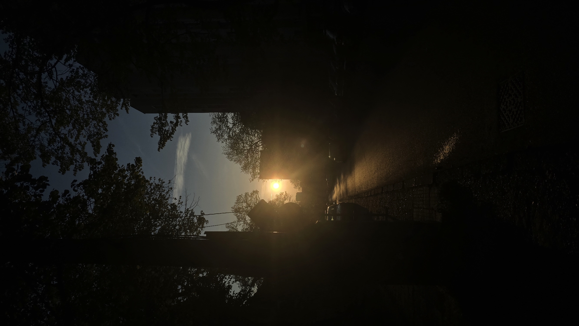The image, likely taken at ground level, depicts a sunlit street scene in Stara Zagora, Bulgaria. The view is partially obstructed by the silhouettes of trees and buildings on either side, framing a central view of the setting sun. The sun is positioned near the center of the frame. A street runs towards the sun, with some parked vehicles visible. The roadway appears wet, reflecting the sunlight. The sky is visible between the trees, showing a gradient from blue to light yellow. A building to the right has a small window. The overall scene has a strong contrast between dark shadows and bright sunlight.