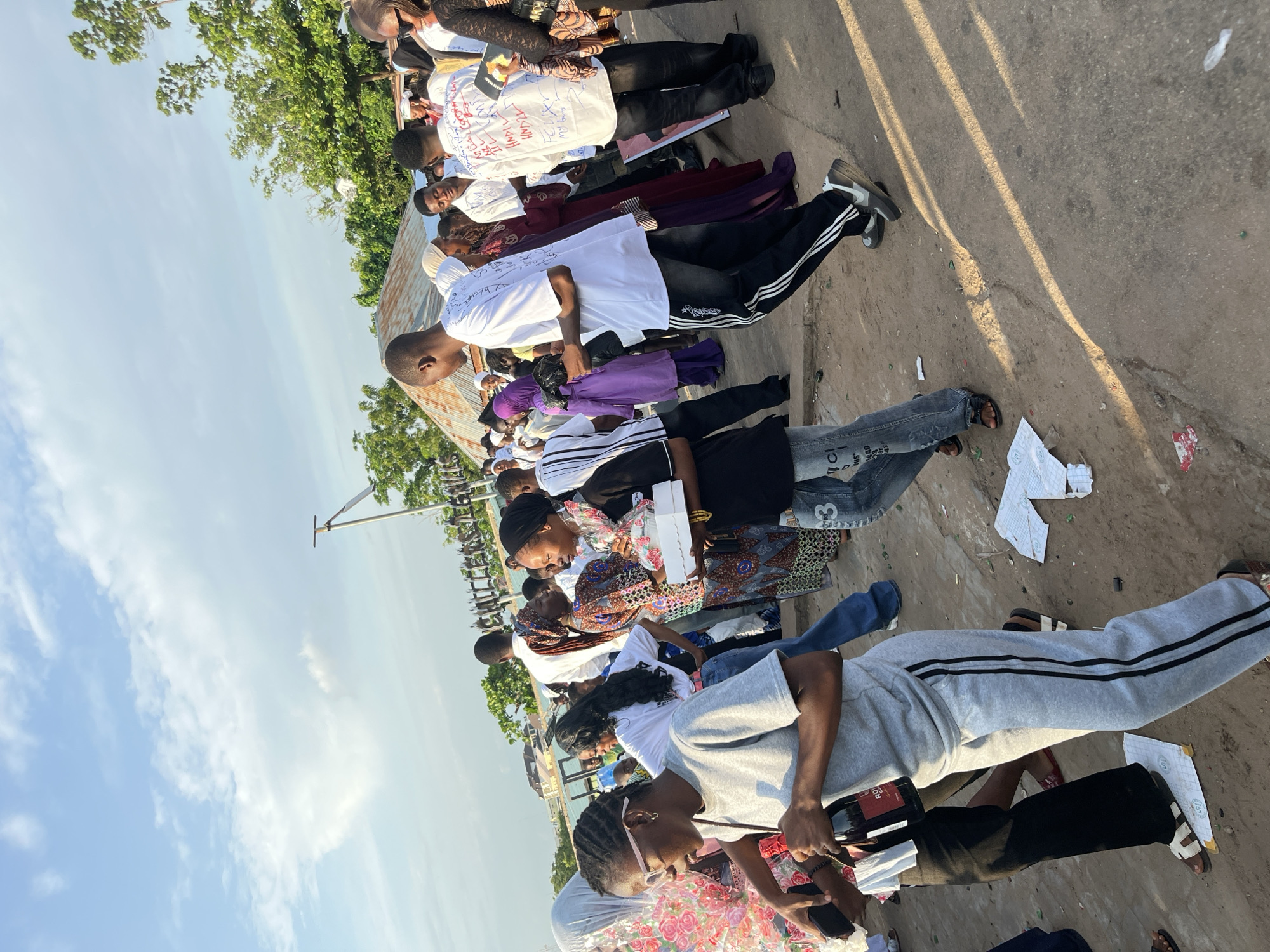 In a sun-drenched outdoor scene, a crowd of people, predominantly dressed in casual and traditional attire, are gathered on a dusty ground. The sky above is a pale blue with scattered white clouds. Several individuals are seen interacting with each other, some carrying gifts or food items. A woman in the foreground is wearing a grey tracksuit and glasses, holding a bottle and a phone. To her right, another woman in a black hijab and patterned dress is holding a white box. In the background, a corrugated metal roof structure is visible, along with trees and a body of water. The ground is littered with debris, including papers and bottles. The overall atmosphere appears to be that of a festive or celebratory event.
