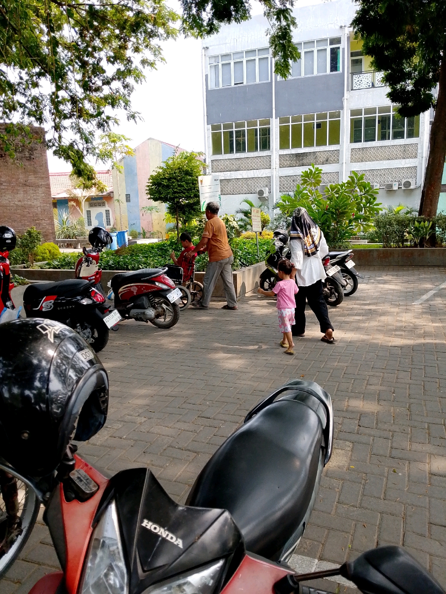 A full-shot, eye-level, outdoor photograph captures a paved parking area adjacent to a building in Kediri City, Indonesia. In the foreground, the seat and handlebars of a red and black motorcycle are visible. Several other motorcycles are parked behind it, including a black scooter with a white "3" logo and a red scooter. To the left, a dark brick building is partially visible behind a tree with green foliage. A light-colored building with multiple windows and air conditioning units stands in the background. In the midground, a man in an orange shirt and gray pants pushes a young boy in a wheelchair. A woman wearing a black hijab and white shirt, and black pants walks with a young girl in a pink shirt and patterned shorts. The ground is covered with interlocking gray pavers.