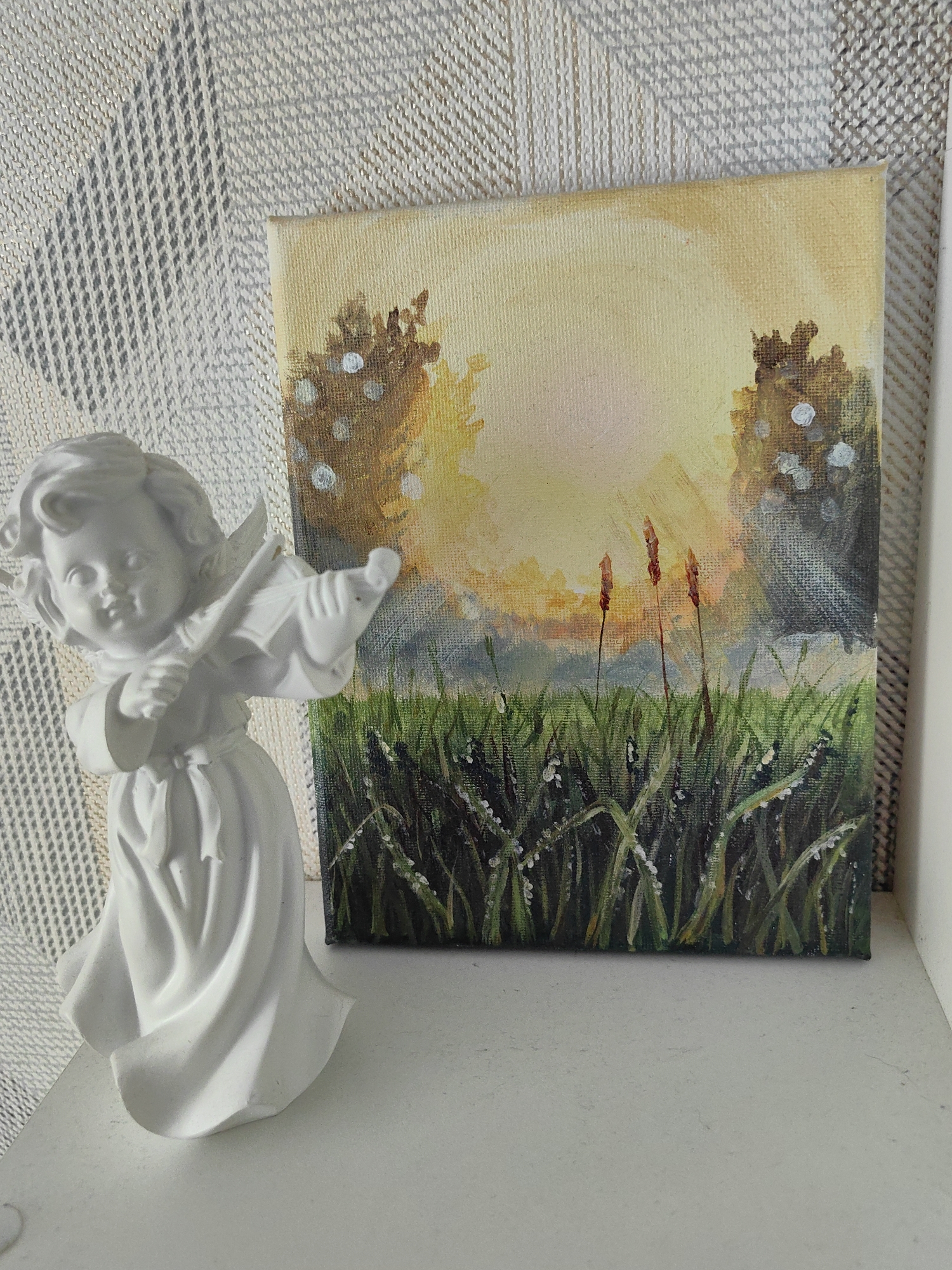 A white statuette of a cherub playing a violin stands to the left of a small canvas painting. The painting depicts a bright, hazy sunset over a field of tall grass, with a hint of trees on either side. The cherub is facing the painting, with its violin held at a playing position. The background behind the cherub is a textured fabric. The objects are placed on a white surface, likely a shelf or table. There is no indication of a specific location in Ufa, Russia.
