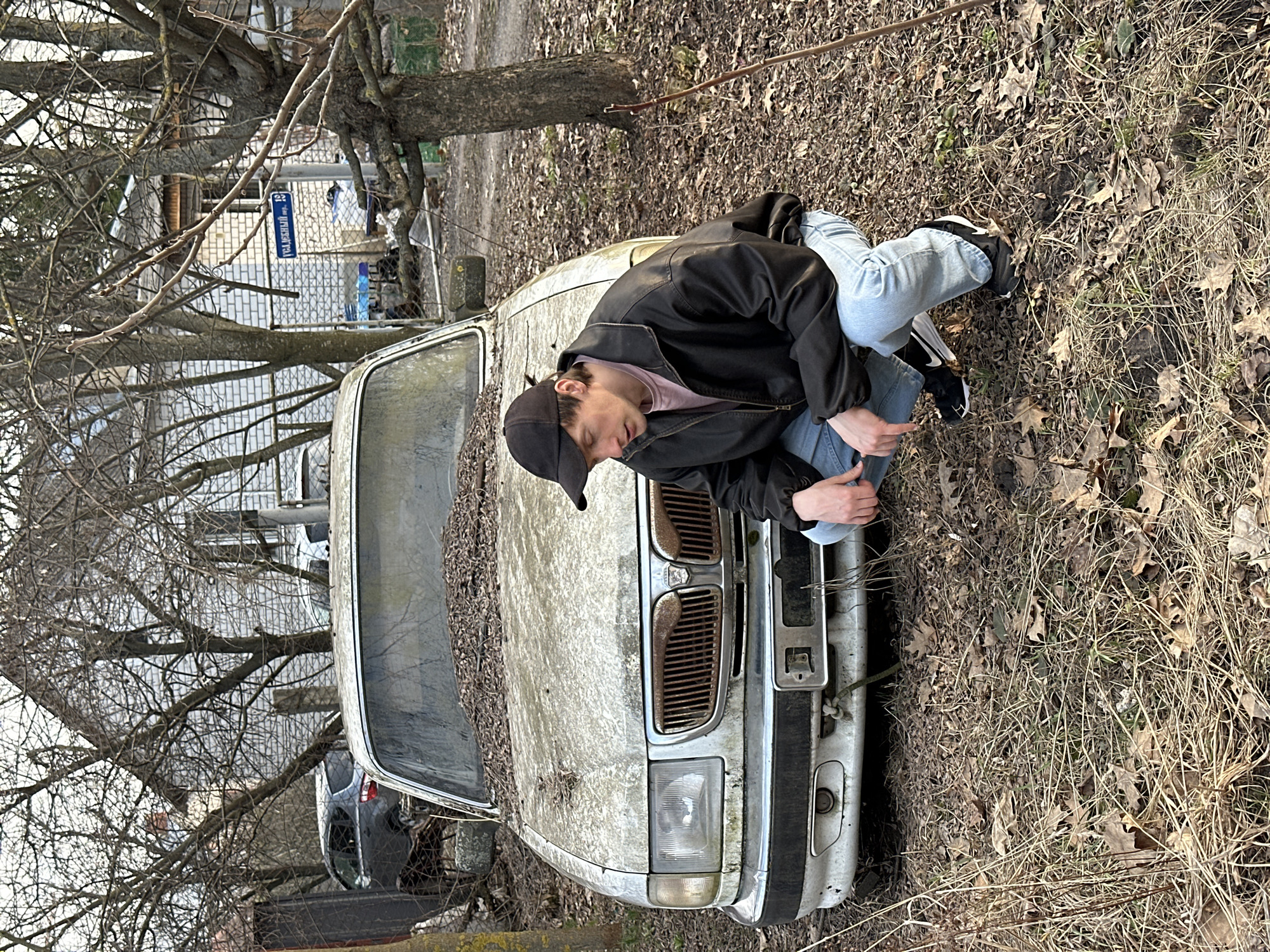 A young man is squatting on the hood of a dilapidated, dust-covered silver car, possibly a vintage model, surrounded by dry leaves and grass. He is wearing a black baseball cap, a black jacket, light blue jeans, and black sneakers with white soles. The scene appears to be an abandoned or neglected area. In the background, bare branches of trees obscure a view of buildings and a street sign that reads "Садовый пр-д, 14" (Sadovy proyezd, 14). Based on the address, the location is in Kursk, Russia. A portion of a dark-colored car is visible behind the silver one.