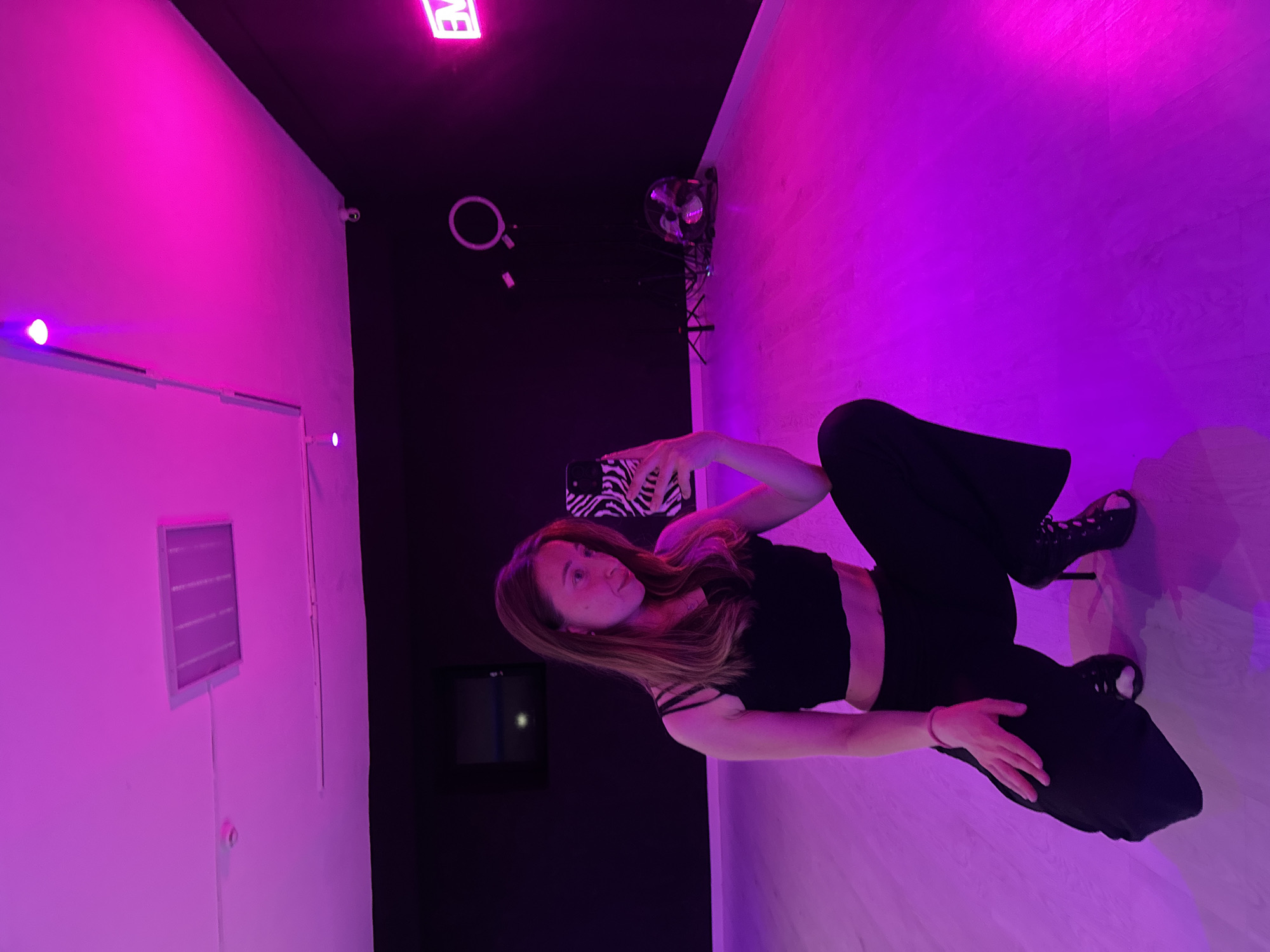 A young woman wearing a black top and pants is posing for a selfie in a room illuminated with pink and purple lights. She is holding a phone with a zebra-striped case. The room has a black wall with a ring light and a fan. There is a white wall with a vent, and a black framed object with a rectangle of light in the black frame. The room is likely a dance or photography studio. Based on the information provided, there is no indication this photo was taken in Chaikovsky, Russia.