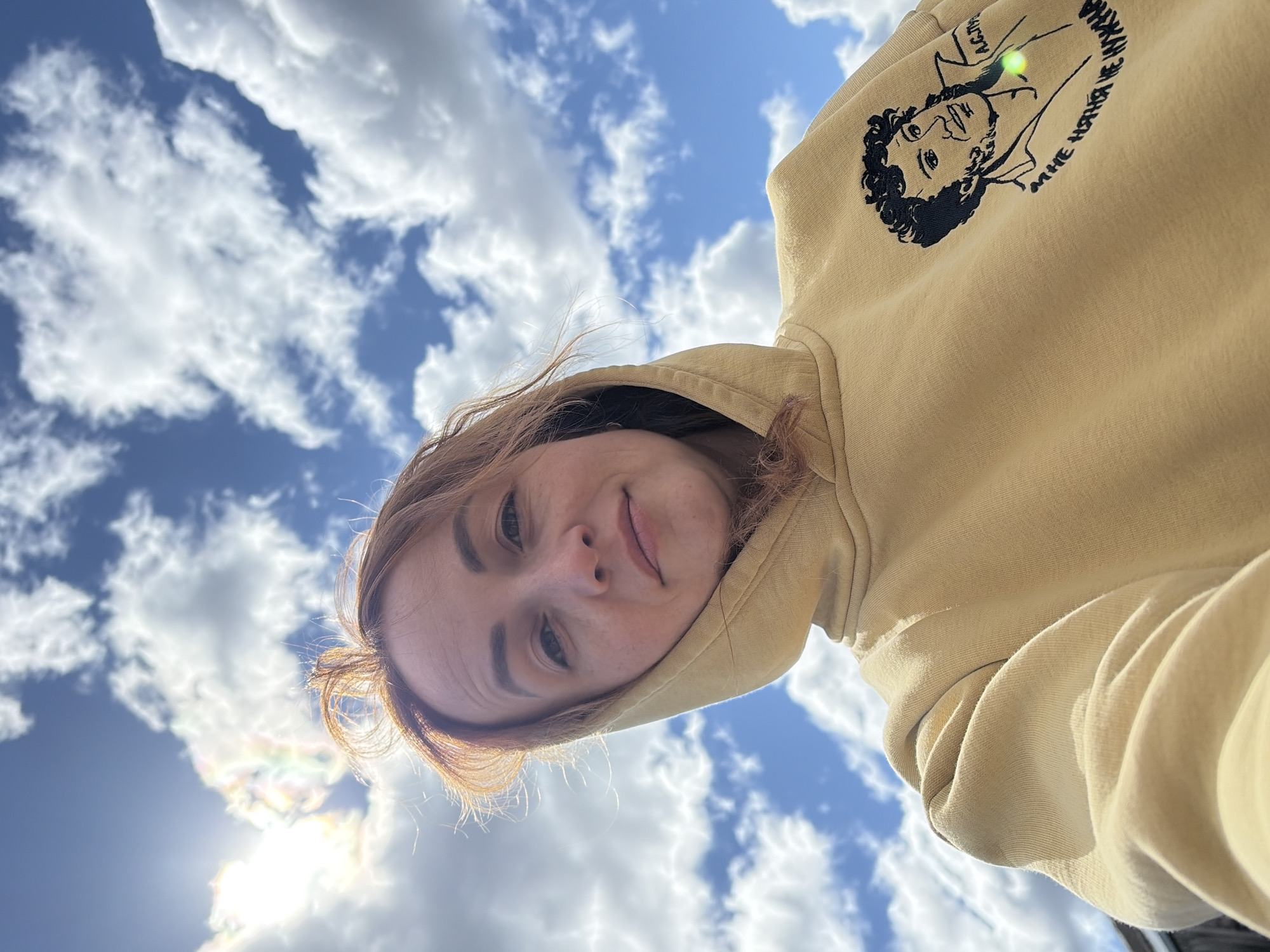 The image shows a woman with light brown hair wearing a yellow hoodie with a graphic on the upper right side. The graphic appears to be a line drawing of a person with curly hair, possibly a portrait. The woman is looking down at the camera with a slight smile. The background is a bright blue sky with scattered white clouds. The sun is visible in the lower left corner, creating a lens flare.
There is no indication of the location being Chaikovsky, Russia, or any other location.