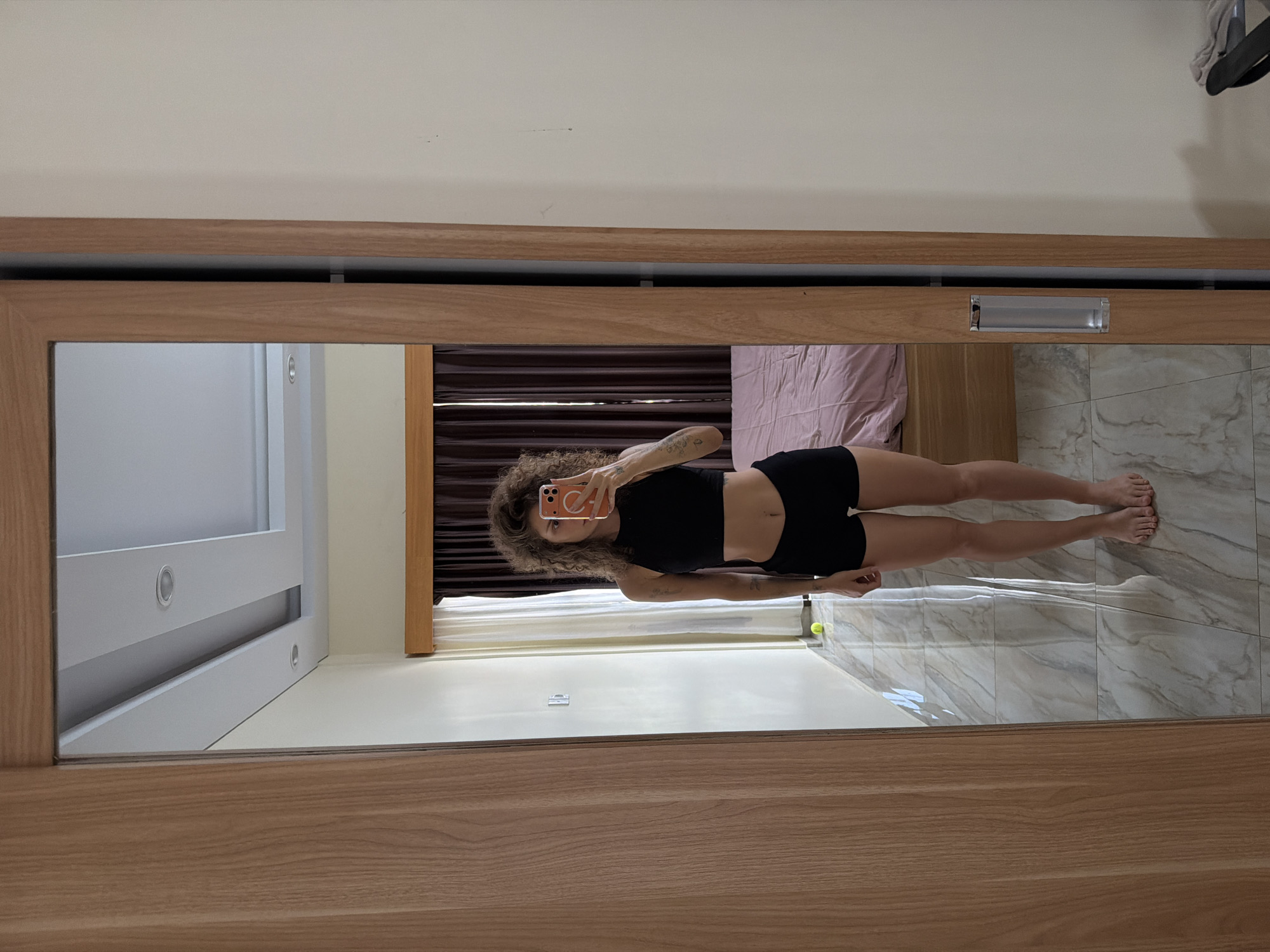 A full-length mirror selfie captures a person with voluminous curly brown hair, wearing a black sleeveless crop top and black athletic shorts. The individual is holding a smartphone with an orange or reddish case, extending their right arm to take the photograph. A tattoo is visible on their right arm.
The mirror is set within a light-colored wooden frame, possibly part of a sliding wardrobe door, featuring a horizontal silver handle. The reflection reveals a modern indoor setting, likely a bedroom. In the background, there's a light cream-colored wall, a bed with a light pink sheet or duvet, and dark brown textured blinds or curtains. The floor is covered with light-toned, possibly marble or tiled, material featuring subtle gray veining. A light-colored wooden furniture piece, resembling a bedside table or dresser, is also visible.
Behind the person's reflected figure, a white sliding door with circular recessed lights or handles can be seen, indicating a closet or another room feature. A yellow-green tennis ball is present on the tiled floor near the person's left foot in the reflection, and a hanger with clothing items is visible in the upper right corner of the reflection. The scene appears to be well-lit, suggesting daytime. No discernible text is visible within the image. The implied location is a private residence in North Kuta, Indonesia.
