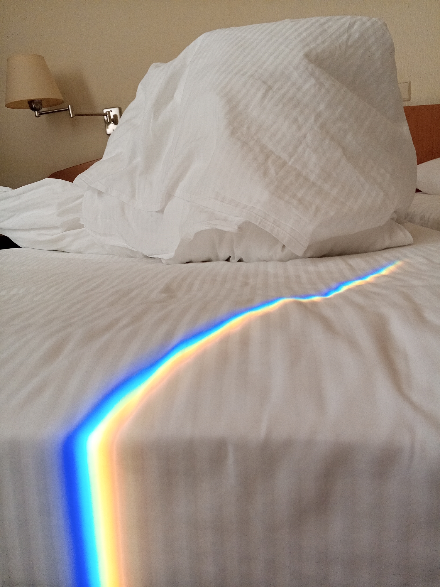 The image depicts a rumpled white bed in a room with a wall-mounted lamp. A prism of light, exhibiting the full spectrum of colors (blue, yellow, orange), is projected onto the bedspread. This is not Saint Petersburg, Russia.
