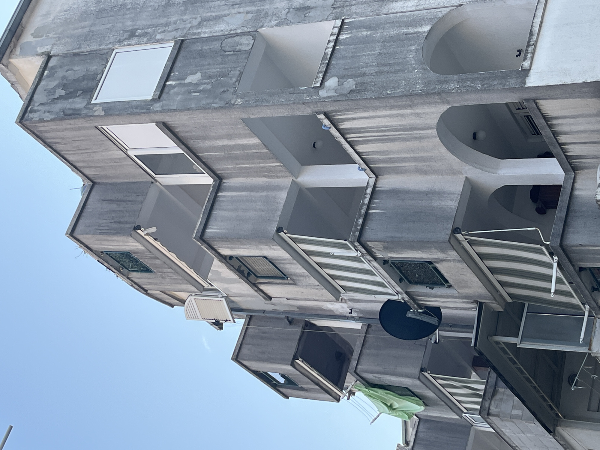 This is a low-angle, full-shot photograph of the side of a weathered, concrete apartment building, with the bright blue sky as the backdrop. The building appears to be in a state of disrepair, with visible staining and peeling paint on its exterior. The structure is characterized by numerous balconies and windows, many of which are recessed or have an angular design. Some of these openings are empty, revealing the dark interiors of the apartments, while others contain modern windows.
Several balconies are equipped with retractable awnings, some of which are striped in green and white, providing shade. Other notable objects include what appears to be an air conditioning unit on one balcony, a dark satellite dish, and some laundry hanging on a clothesline, including a bright green garment. The overall impression is one of a residential building in a sunny, possibly Mediterranean location, given the context of Budva, Montenegro. The time of day appears to be daytime, as indicated by the bright sky. There is no visible text in the image. The scene evokes a sense of everyday life in a residential setting, with elements of both wear and modernity.