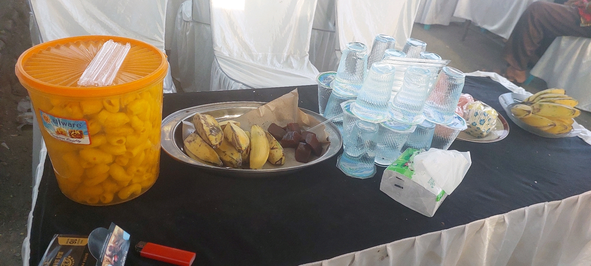 In Bukur, Indonesia, a black tablecloth covers a table laden with food and drinks. On the left, a large orange plastic container filled with macaroni is prominently displayed. Adjacent to it, a silver platter holds a bunch of bananas and some cubed dark-colored food, possibly dates. To the right, a stack of clear plastic cups is arranged, with a package of napkins placed nearby. Further to the right, another platter contains more bananas. The background features white-covered chairs, suggesting an event or gathering. A person in dark brown clothing is partially visible in the upper right corner, seated. A red lighter and a pack of cigarettes are on the table in the lower left corner.