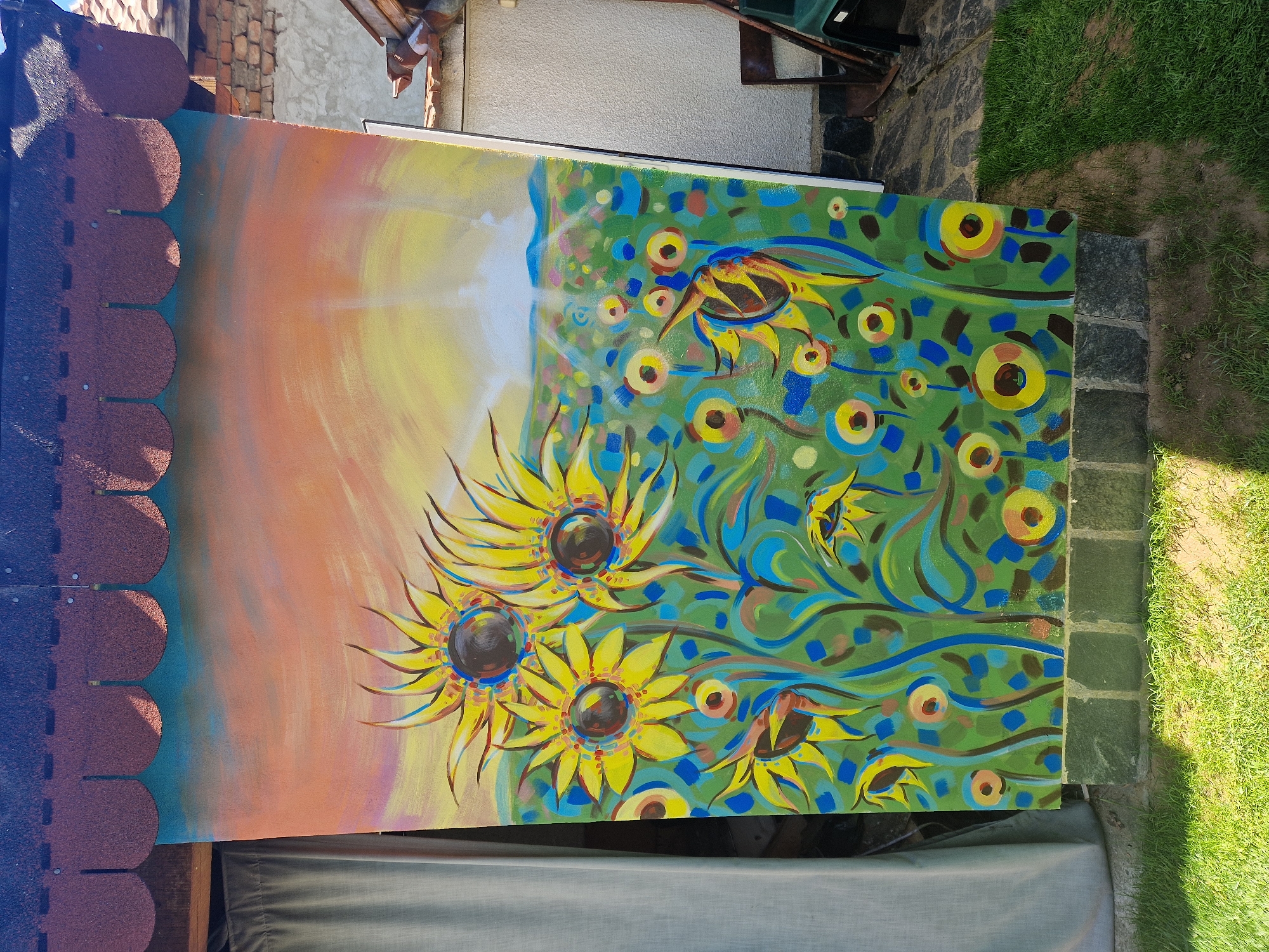 The image shows an oil painting of sunflowers leaning against a structure outdoors. The painting is divided into two distinct sections. The left side features a warm sunset sky, transitioning from orange to yellow. The right side displays a stylized green background with sunflowers interspersed with blue and brown circles. The sunflowers themselves are painted with bright yellow petals and dark brown centers.
The painting is positioned outside, resting against a roof section with scalloped roofing tiles. Behind the roof, a brick wall is visible. The bottom of the painting rests on a grey, likely fabric-covered surface. To the right of the painting, there's a paved stone area transitioning into a grassy lawn. A green garden tool, possibly a rake, is partially visible in the top right corner.