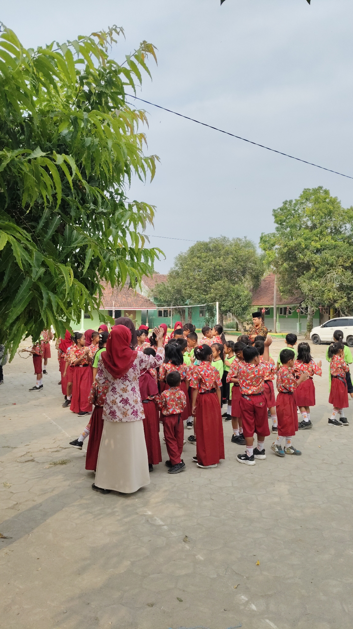 In Banjarsari, Indonesia, a group of young students, predominantly girls, stand in a courtyard. They are wearing matching red and white patterned shirts and red skirts or shorts, suggesting a school uniform. One adult woman, wearing a maroon headscarf and a floral patterned blouse with a long, cream-colored skirt, stands at the forefront, facing away from the camera, seemingly addressing the children. Another adult, a man in a military-style uniform, is visible in the background, also facing the students. The children are arranged in a loose formation, with some looking towards the adults and others interacting with each other. The environment appears to be an outdoor school setting, with a paved courtyard, a low building with a red tiled roof in the background, and lush green trees framing the scene. A volleyball net is visible in the middle ground, and a white car is parked on the right side of the frame. The sky is overcast.