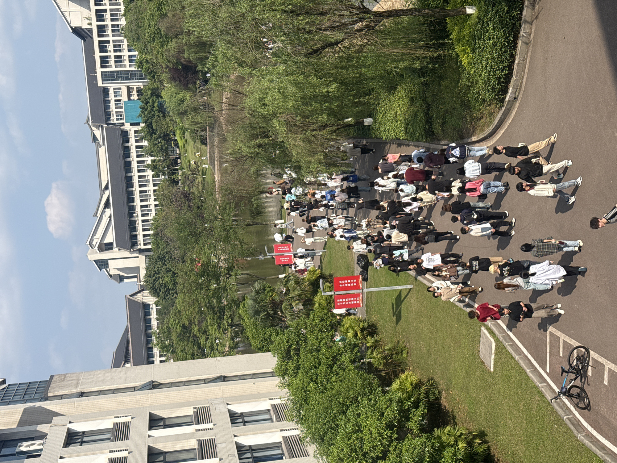 A high-angle, outdoor daytime shot shows a group of people lying down on a paved path next to a grassy area and a body of water, likely a lake or pond. The location is likely near a university campus in Jiangjin, China, as evidenced by the architectural style of the surrounding buildings.
The paved path curves slightly, and the people are lying in a row, close to each other. Most of them appear to be young adults, possibly students. The majority are dressed in casual clothing such as t-shirts, sweaters, and pants.
Adjacent to the path is a grassy area with trees and bushes. There are also several red signs with white writing on them. The buildings in the background appear to be university dormitories or academic buildings, with a multi-story construction and multiple windows. A bicycle is parked on the side of the path.