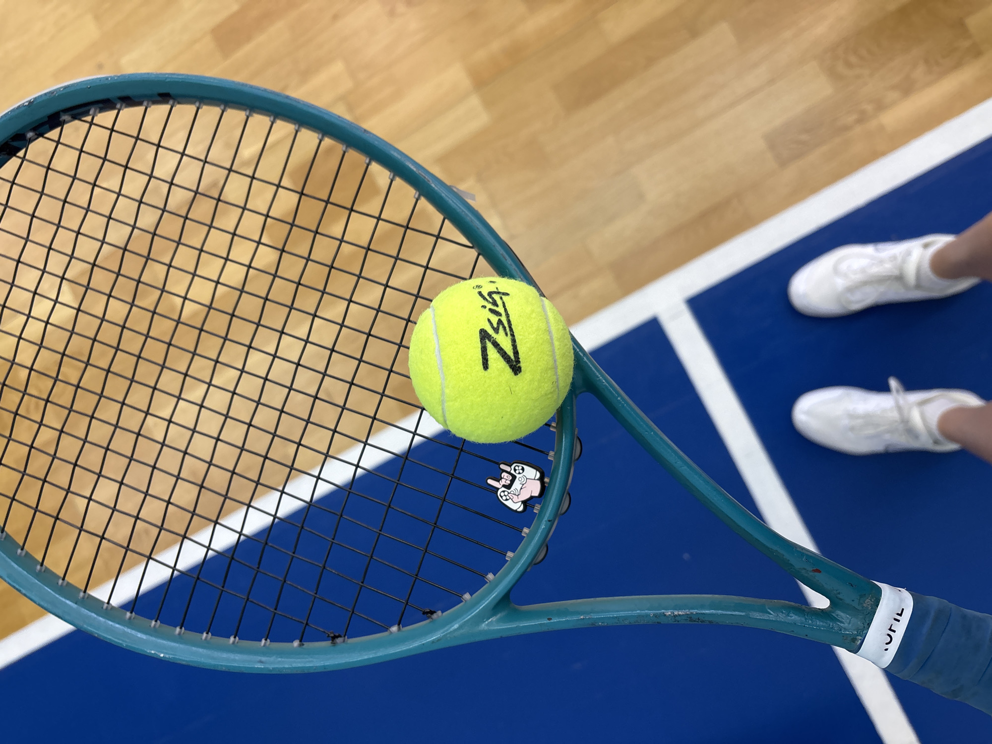 A tennis racket and a yellow tennis ball are positioned on a blue and white tennis court, next to a wooden floor. A pair of white sneakers are partially visible on the right side of the image. The racket is light blue, with black strings, and a "ZSig" tennis ball rests in the center of the racket's strings. The location is the city of Chaikovsky, Russia.