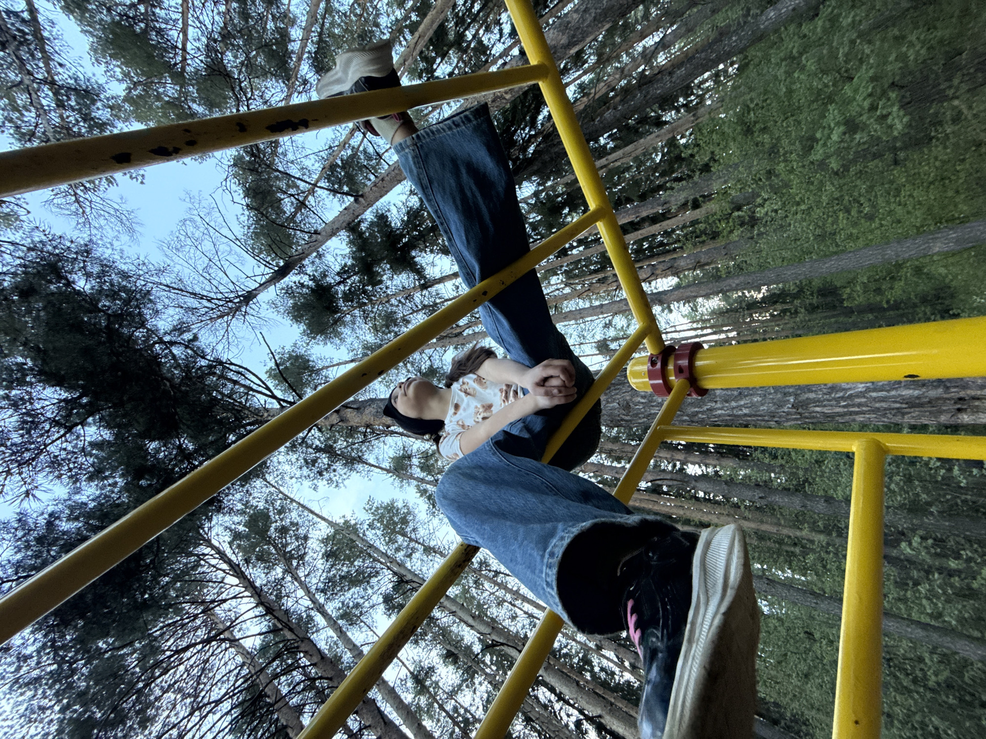 A person, lying on yellow metal monkey bars, is photographed from a low angle against a background of tall, thin trees. The person wears blue jeans, a light-colored t-shirt with a pattern, and dark shoes. Their head is tilted back, with their hands clasped. The scene includes metal bars that are painted yellow and that make up the monkey bars structure. The background includes tall, thin trees, possibly in a forest or park in Votkinsk, Russia.