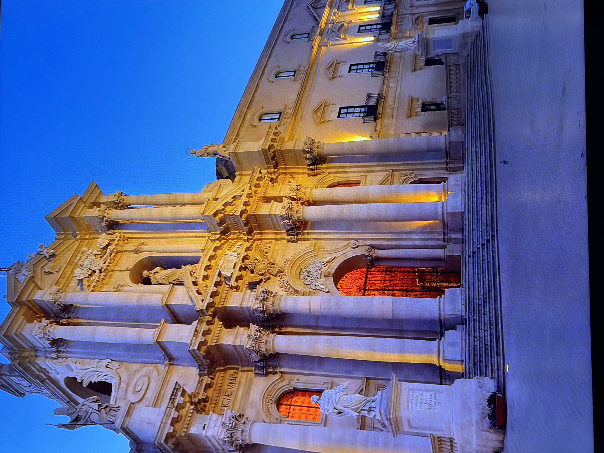 The image depicts a grand, ornate building, likely a cathedral or a significant historical structure, illuminated by warm, artificial light against a deep blue twilight sky. The architecture features classical elements such as columns, pediments, and intricate carvings. Statues are visible on pedestals and within niches on the facade. A large, arched doorway with decorative metalwork is centrally located. Stairs lead up to the entrance, and the ground in front appears to be a paved plaza. There is a white vehicle parked to the right of the building. No people are clearly visible in the scene. The location of Chirchik, Uzbekistan is not evident from the visual information presented.