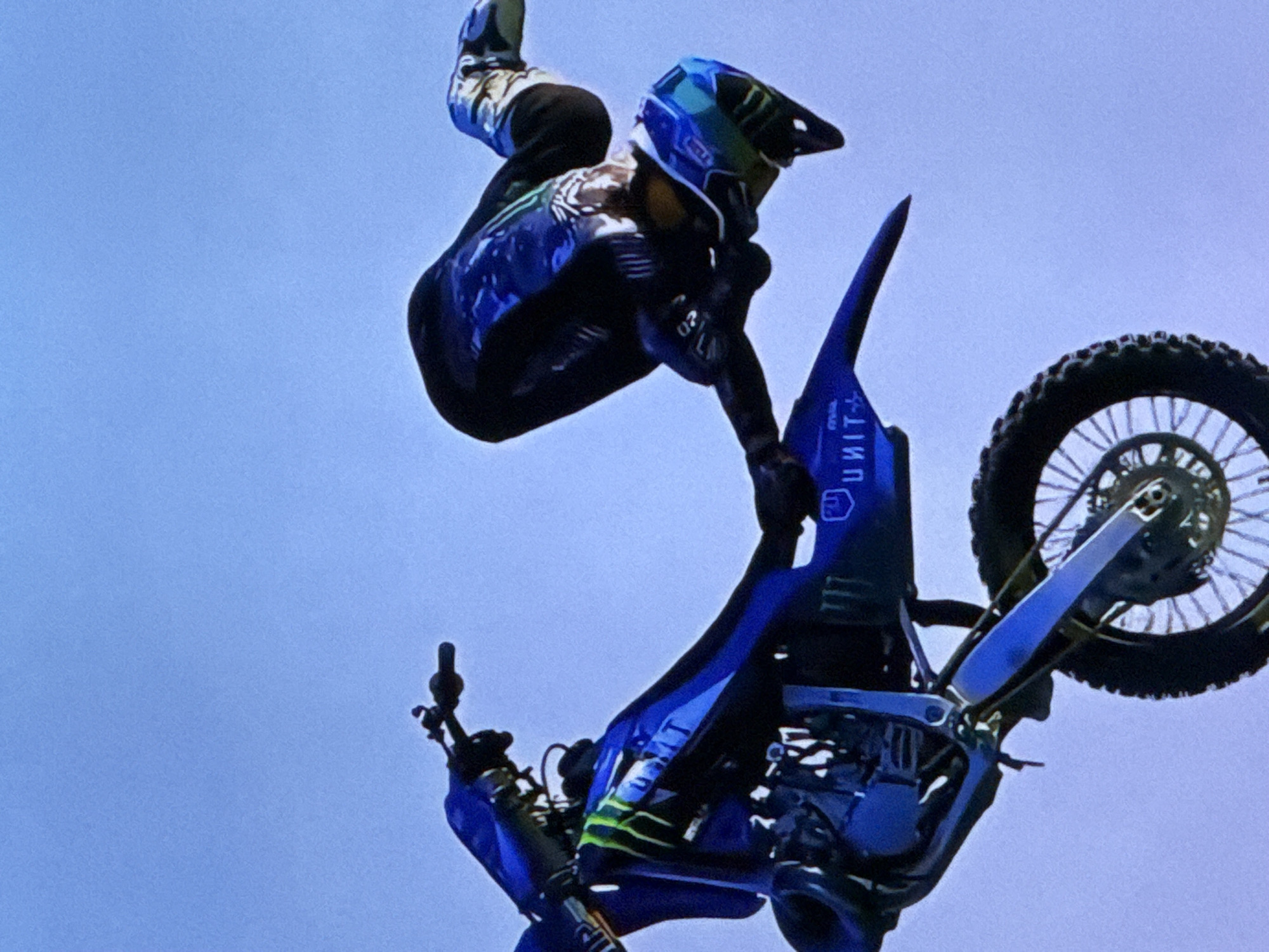 The image depicts a motorcyclist performing a mid-air stunt on a blue dirt bike against a clear blue sky. The rider is wearing a black and blue racing suit and a helmet. The motorcycle has prominent green and white graphics. The stunt appears to be a backflip, with the rider and bike inverted. The scene is captured from a low angle, emphasizing the height of the jump. There is no visible urban environment or indication of the city of Cahul, Moldova. The image is a still photograph with no discernible action or interaction beyond the described stunt.