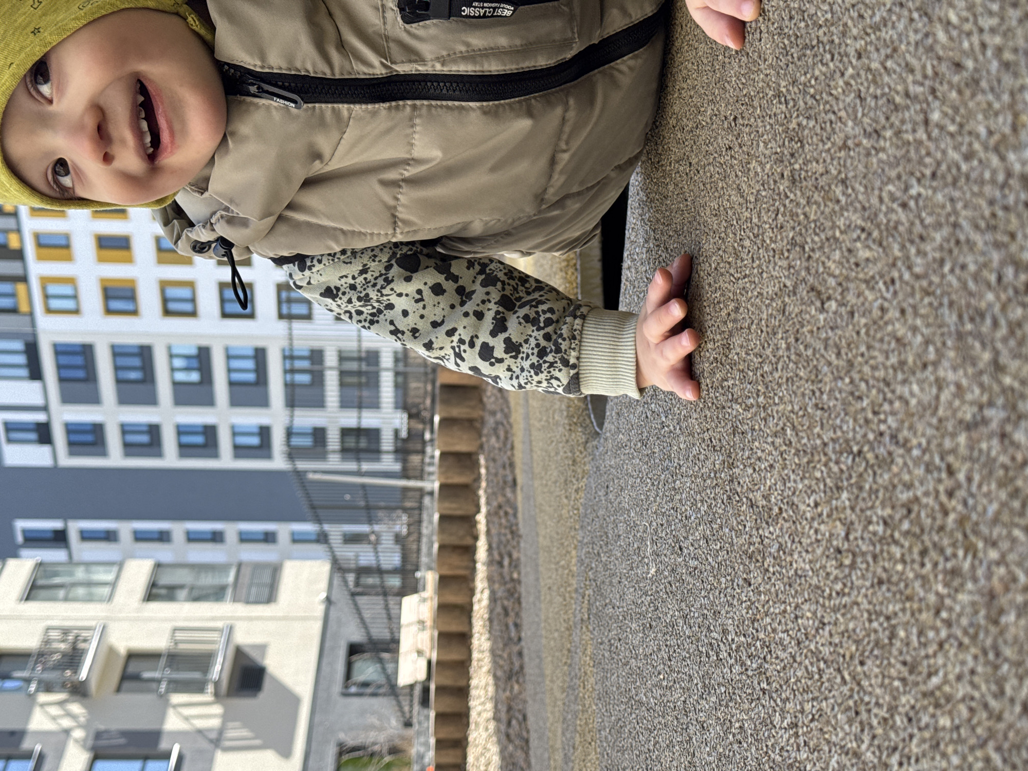 A child with a yellow beanie and a tan puffer vest is seen in the foreground, with the modern residential buildings of Izhevsk, Russia in the background. The child's face is visible, smiling. One arm is extended, with fingers resting on a coarse surface. The buildings feature alternating rows of blue and yellow windows, a characteristic design element of newer construction in the city. The setting suggests a playground or recreational area near a residential complex.