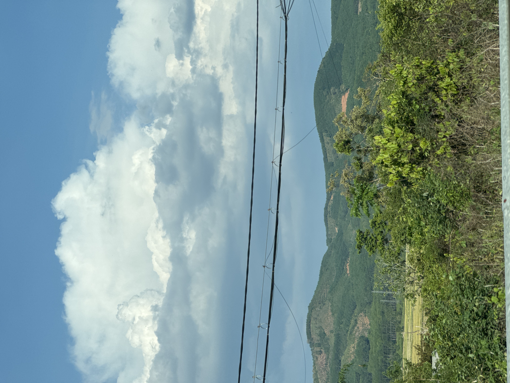 The image depicts a landscape near Đăk La, Vietnam. The foreground contains power lines running vertically, partially obscuring the view. To the left, a sky with white cumulus clouds against a blue background is visible. The right side of the image shows a series of green mountains, with areas of exposed reddish soil visible on some slopes. In the foreground, foliage and trees are interspersed with a possible rice field area.