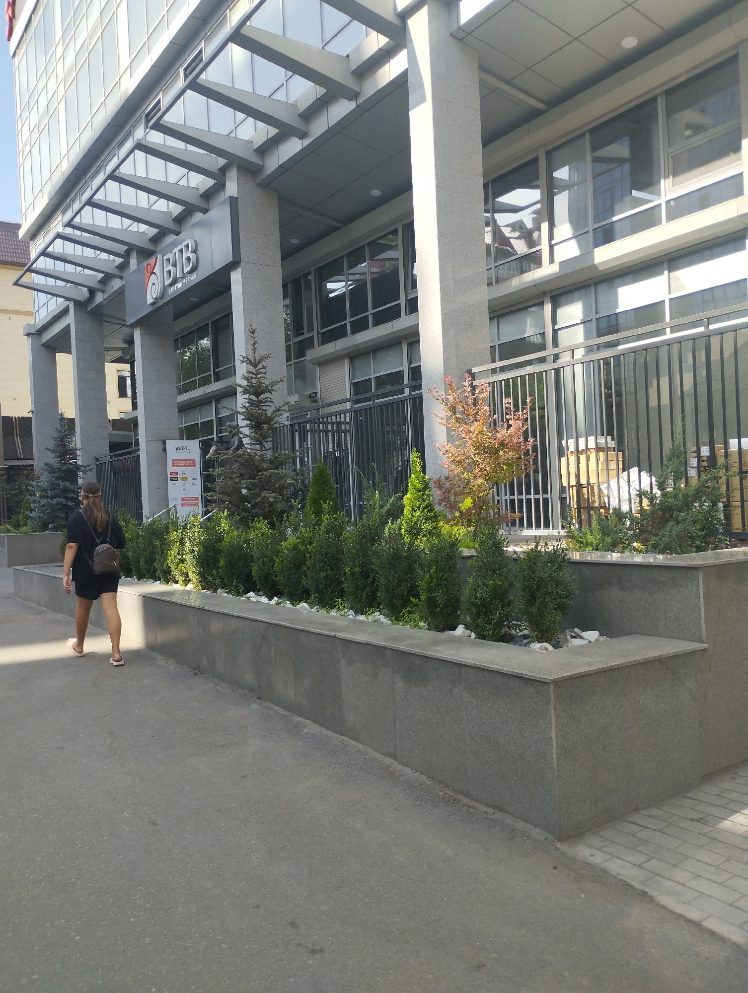In Bishkek City, Kyrgyzstan, a young woman with dark hair pulled back walks away from the camera, wearing a black t-shirt, black shorts, and pink slides. She carries a small brown backpack over her left shoulder. The woman is walking on a paved sidewalk that runs alongside a modern, multi-story building with a glass facade. In front of the building, there is a tiered concrete planter filled with various green shrubs and small trees. A black metal fence separates the planters from the building's entrance area. The building has a prominent sign with the letters "BTB" in red and white. The weather appears to be sunny.