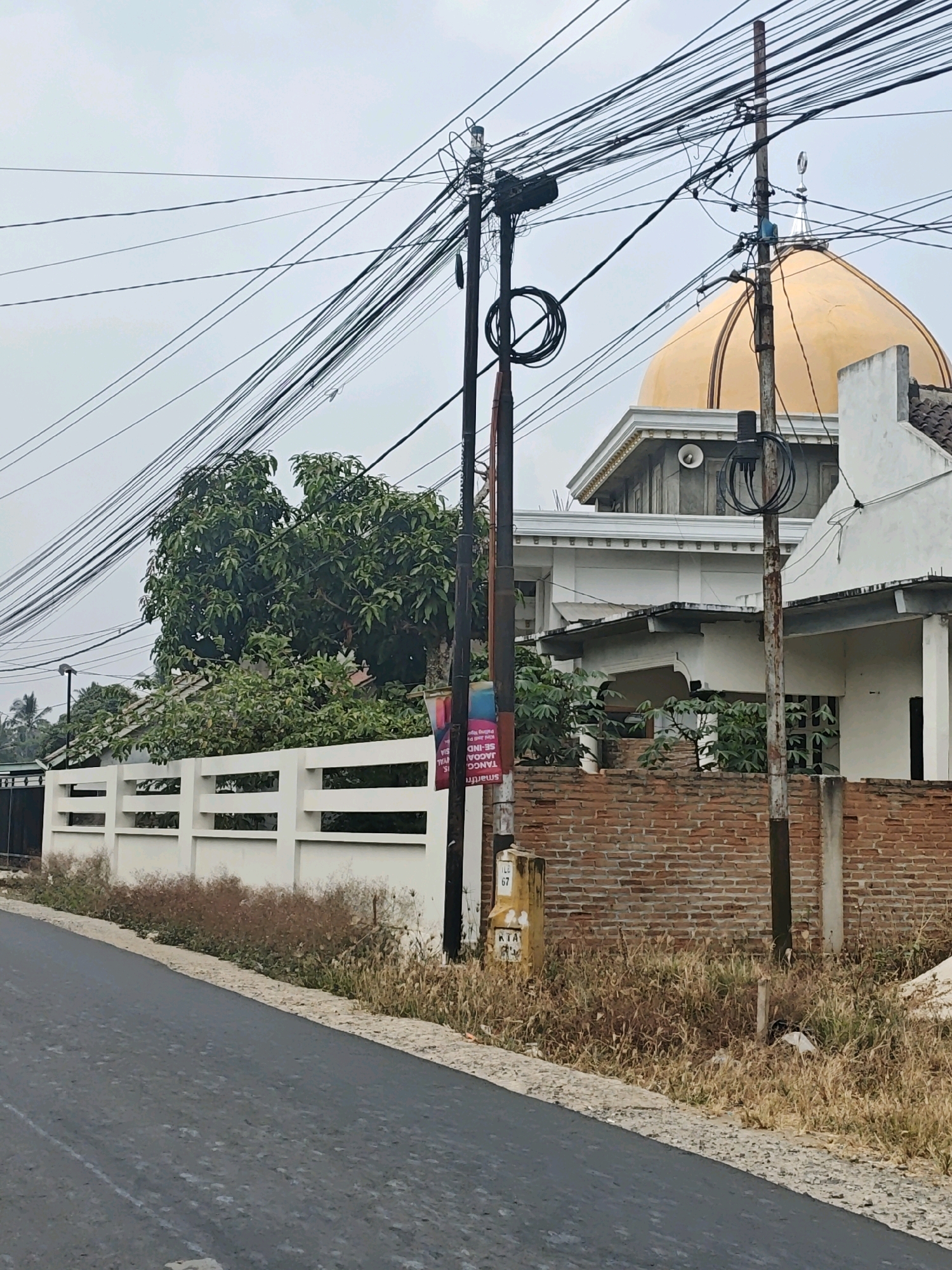 The image depicts a street scene in Sinar Semendo, Indonesia. The foreground shows a paved road with a rough surface and scattered debris. To the right of the road is a dry, overgrown embankment with tall grasses and weeds, bordered by a brick wall. Beyond the brick wall, a white fence runs parallel to the road. In the background, a prominent golden dome of a building, likely a mosque, is visible against a cloudy sky, with a crescent moon atop it. Several utility poles with a dense network of electrical wires crisscross the sky. Adjacent to the mosque is a white building with a covered walkway. Trees with lush green foliage are interspersed with the structures. A colorful poster is attached to one of the utility poles. The overall environment appears to be a residential or mixed-use area with significant overhead wiring.