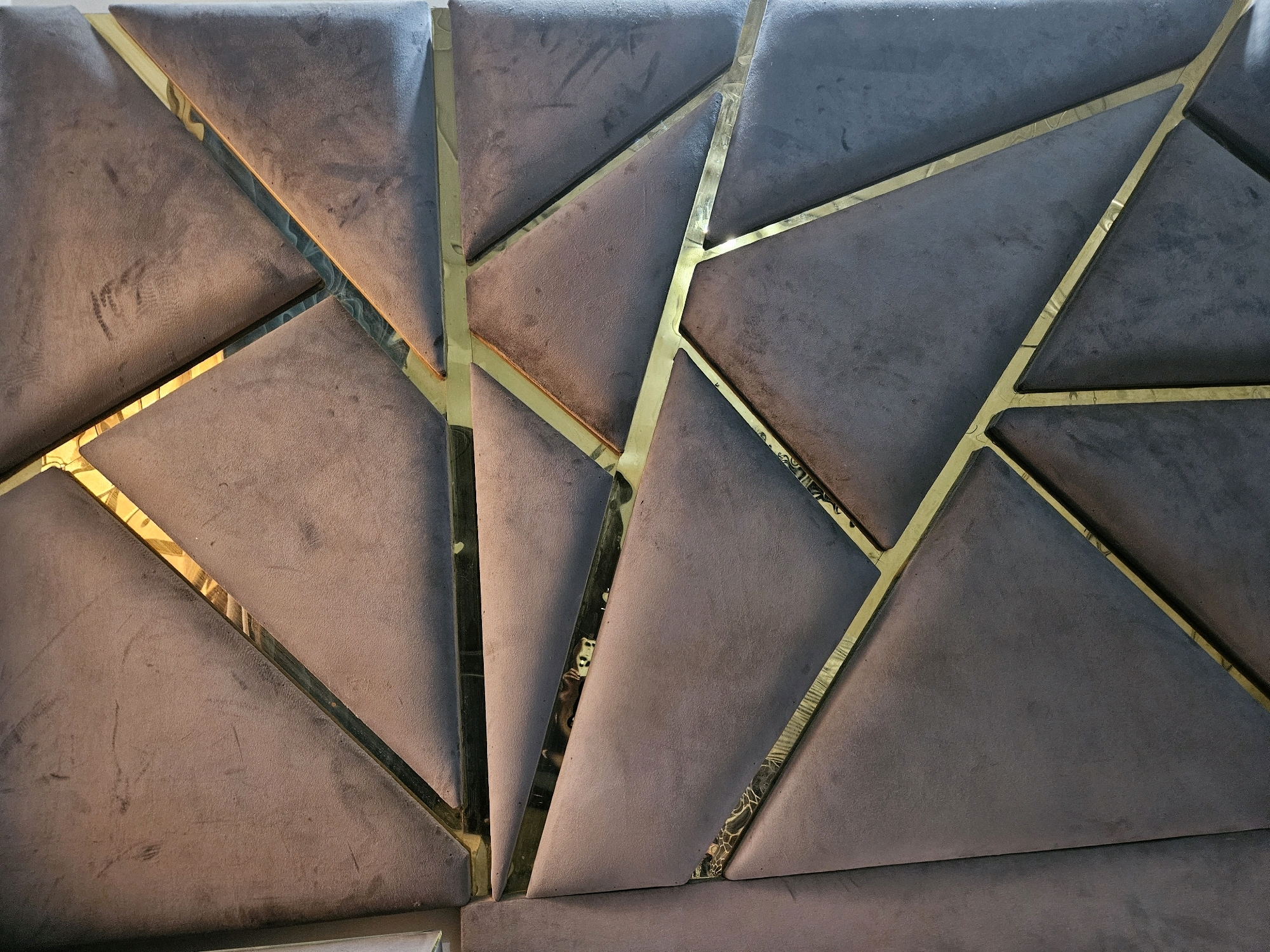 The image displays a close-up view of a padded headboard or wall paneling. The surface is segmented into triangular and polygonal shapes upholstered in a dark grey, velvety fabric. These fabric panels are outlined and separated by thin, metallic gold trim. The gold trim appears to be reflective, with some areas showing distorted reflections of what might be lights or other elements in the environment. The lighting in the scene casts shadows, highlighting the geometric pattern and the texture of the fabric. The overall impression is one of modern, luxurious interior design. This scene is located in Jawad Avenue, Pakistan. There are no people or discernible actions captured in this image; it is a static shot of a decorative surface.