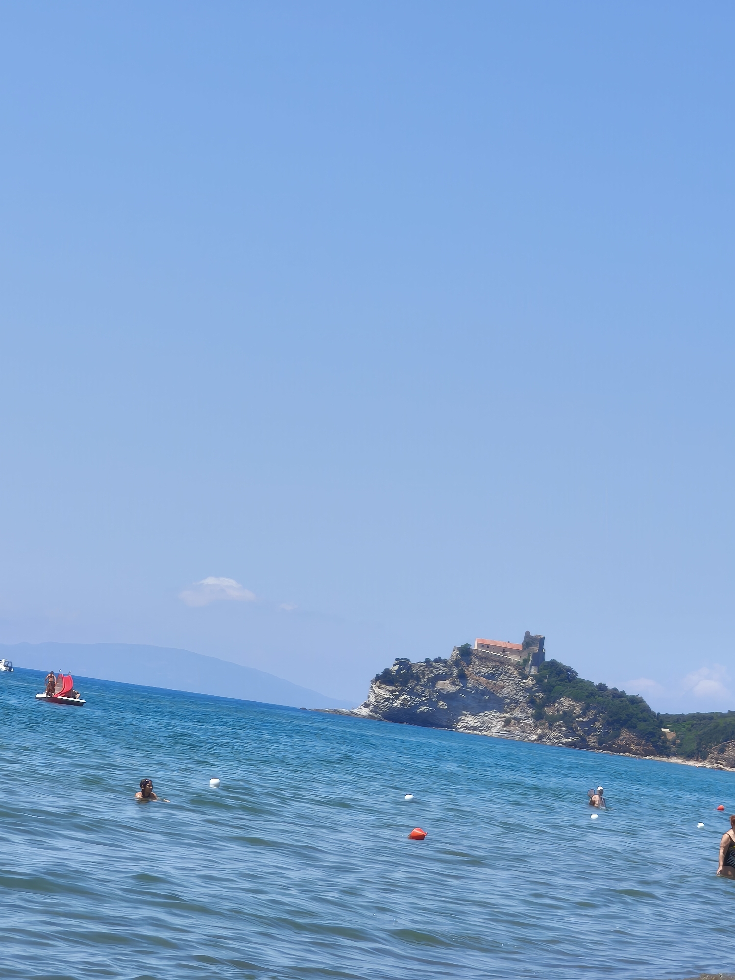 The image depicts a sunny day at a beach in Castiglione della Pescaia, Italy. The clear blue sky and calm turquoise water suggest pleasant weather, likely mid-day. In the foreground, several people are enjoying the sea, with some swimming and others wading near the shore. White buoys and a few orange ones are scattered across the water, marking an area within the sea.
Further out, a small red sailboat with two people on board is visible. In the middle ground, a rocky island rises from the sea, crowned by an old stone building, possibly a fortress or castle, with a reddish-tiled roof. The island is covered in lush green vegetation. The distant shoreline shows hills and more greenery under the bright sky.
The overall scene conveys a sense of leisure and relaxation, typical of a summer vacation. There is no visible text in the image. The lighting indicates it is daytime, with the sun casting bright light and creating reflections on the water.