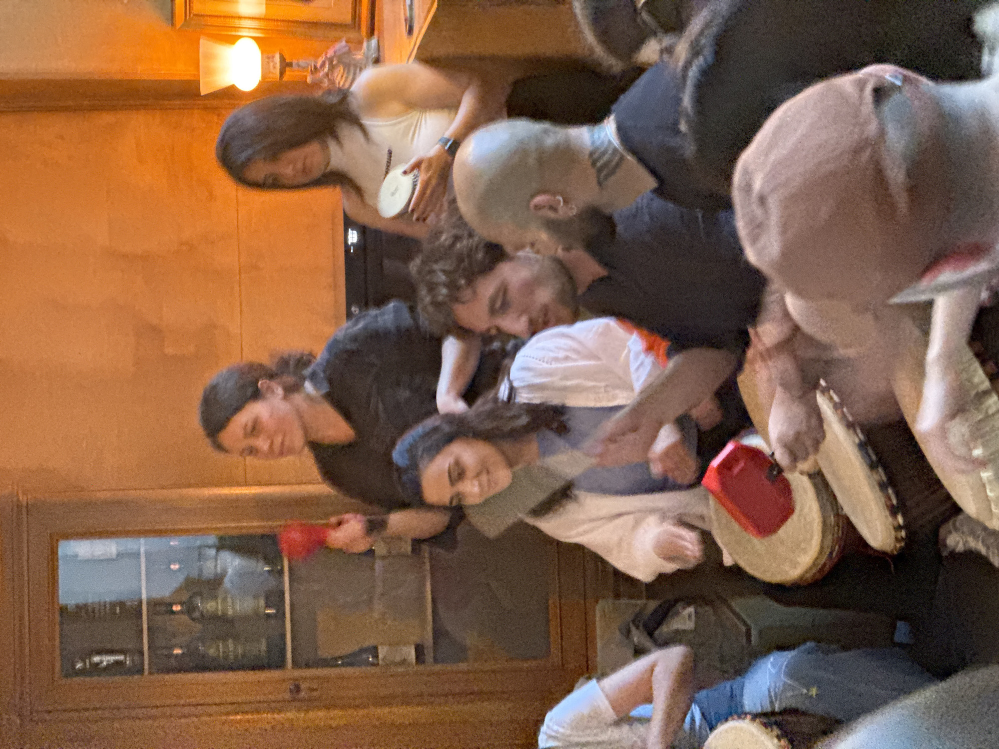 A group of individuals are gathered indoors, likely in a residential setting with wooden floors, walls, and furniture. One person lies prone on the floor. Several people are holding and playing percussion instruments, specifically drums. The lighting is warm and appears to be from artificial sources. The scene appears to be a casual social gathering focused on music-making. The city of Yerevan, Armenia, cannot be verified from the provided imagery.
