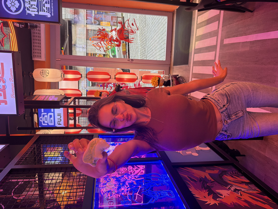 A young woman poses in a brightly lit, Asian-themed space, likely a restaurant. She holds a piece of food up close, puckering her lips towards the camera. She wears glasses, a brown shirt, and light jeans. Behind her, there are rows of red and white lanterns, signs with Japanese writing, and blue-lit artwork. The floor is white with black stripes, mimicking a crosswalk.