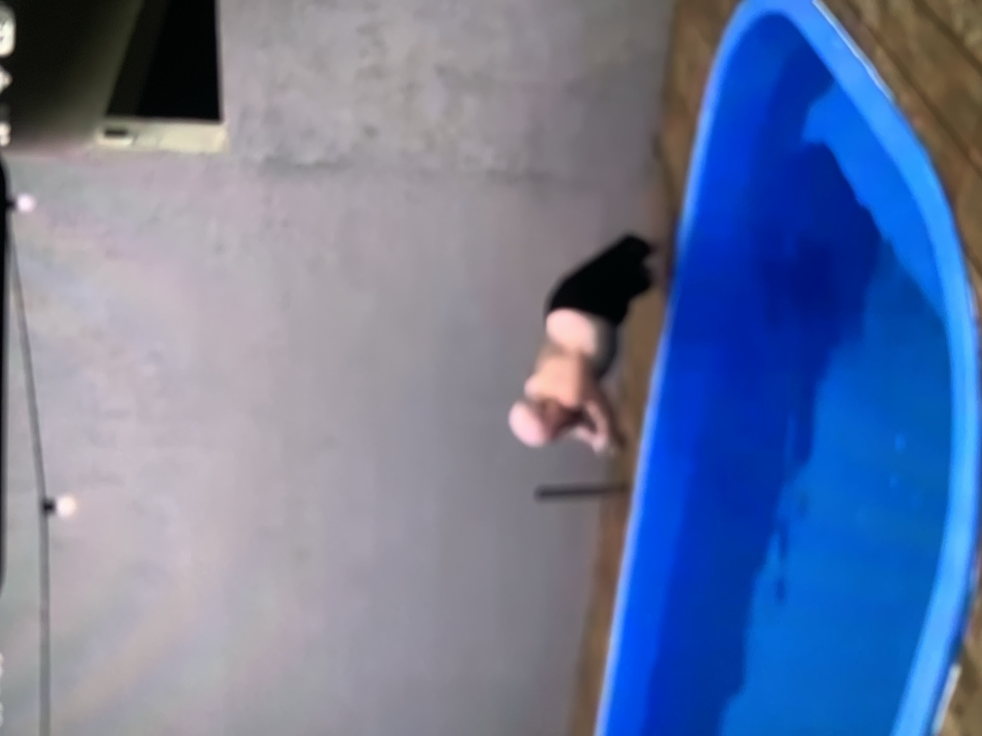 A man is captured mid-air as he dives into a blue inflatable swimming pool. He is balding and appears to be wearing black shorts. The pool is set on a wooden deck, adjacent to a gray wall. Above the scene, a string of lights is visible. The image's low resolution and motion blur make it difficult to ascertain specific details about the man's face or the surrounding environment. No specific location within Stavropol, Russia, can be identified from the provided image.