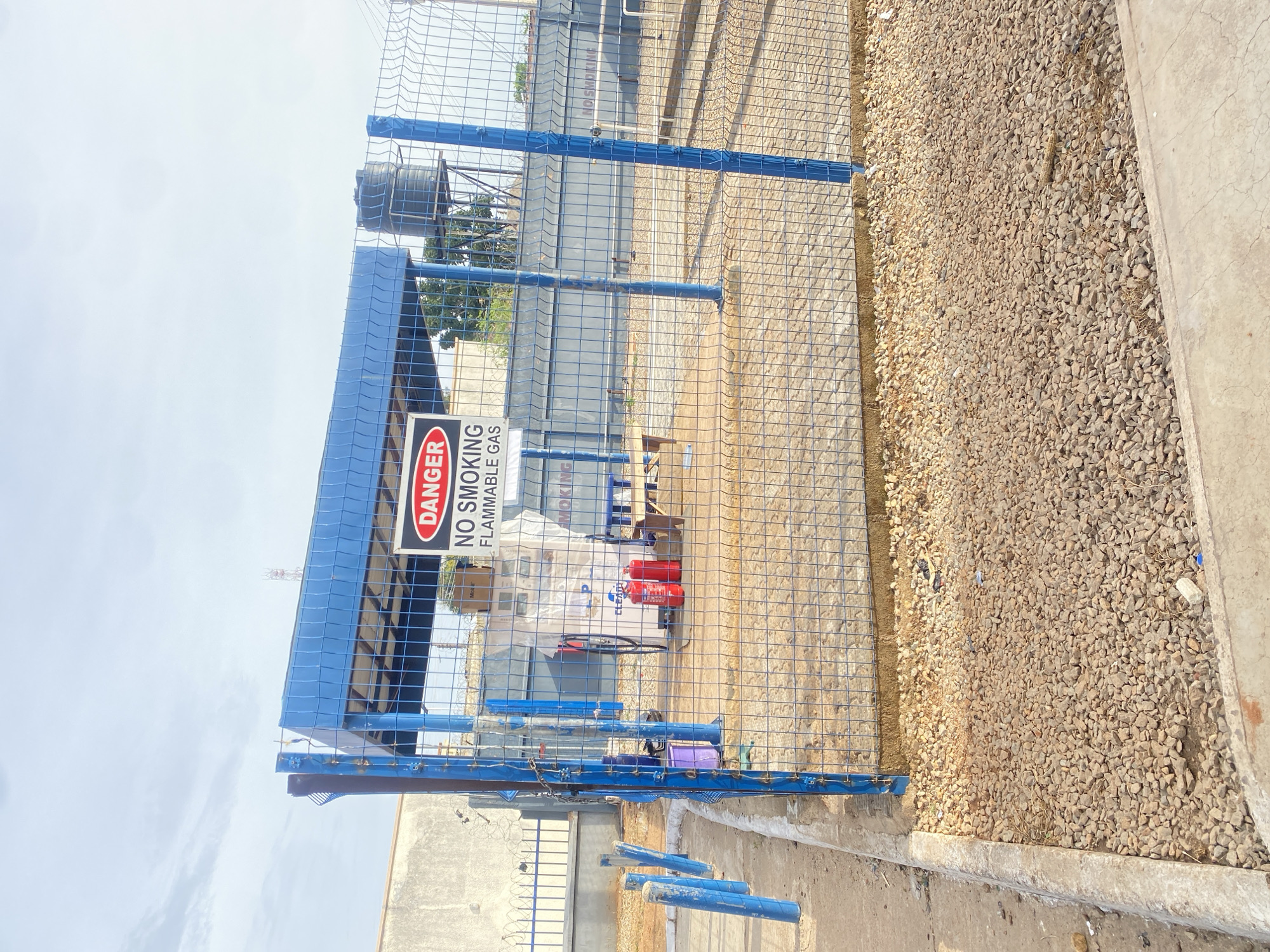 The image shows an outdoor industrial area in Jos, Nigeria. The scene is dominated by a blue metal fence and a blue-roofed structure. A "DANGER NO SMOKING FLAMMABLE GAS" sign is prominently displayed on the fence. Inside the fenced area, there are two red fire extinguishers and what appears to be a bicycle. In the background, a water tank is visible. The ground is covered with gravel and some concrete. The sky is bright, suggesting it is daytime with clear or partly cloudy weather. There are no people visible in the image. The overall impression is one of an industrial safety zone.