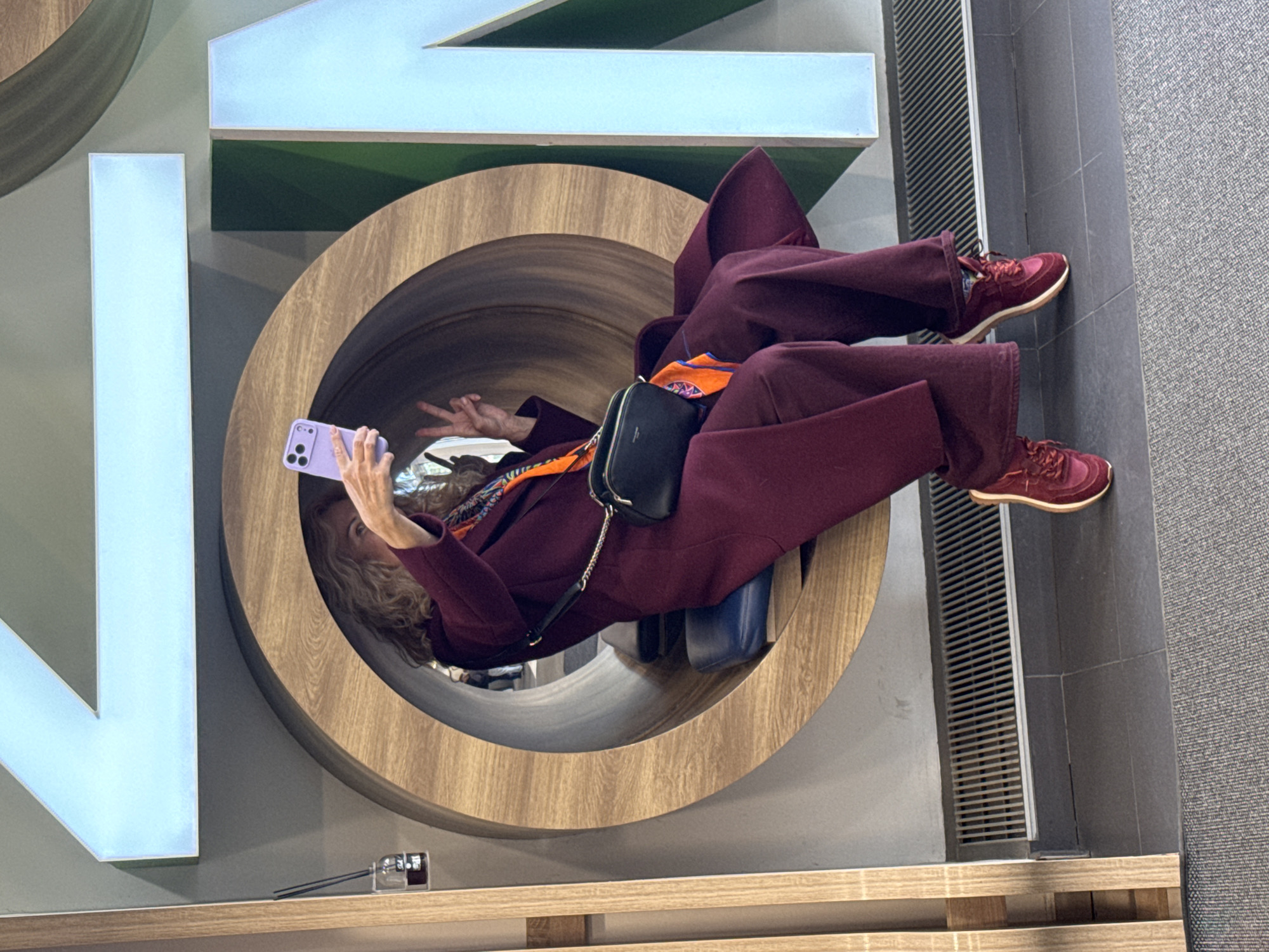 A woman in a maroon pantsuit and matching sneakers is positioned inside a large wooden circular structure, seemingly part of a modern art installation or interactive display. She is holding up a lavender smartphone, possibly taking a selfie or recording a video, and making a peace sign with her left hand. She has a small black purse with a chain strap across her torso, and a patterned orange scarf around her neck. The circular structure is set against a gray wall, with a white and green abstract design visible behind it. There's a wooden shelf or ledge below the structure, and a metallic vent along the wall on the right side of the image. The scene is set in what appears to be a contemporary public space. Based on the overall architectural style and the presence of design elements, the location may be in the city of Saint Petersburg, Russia.
